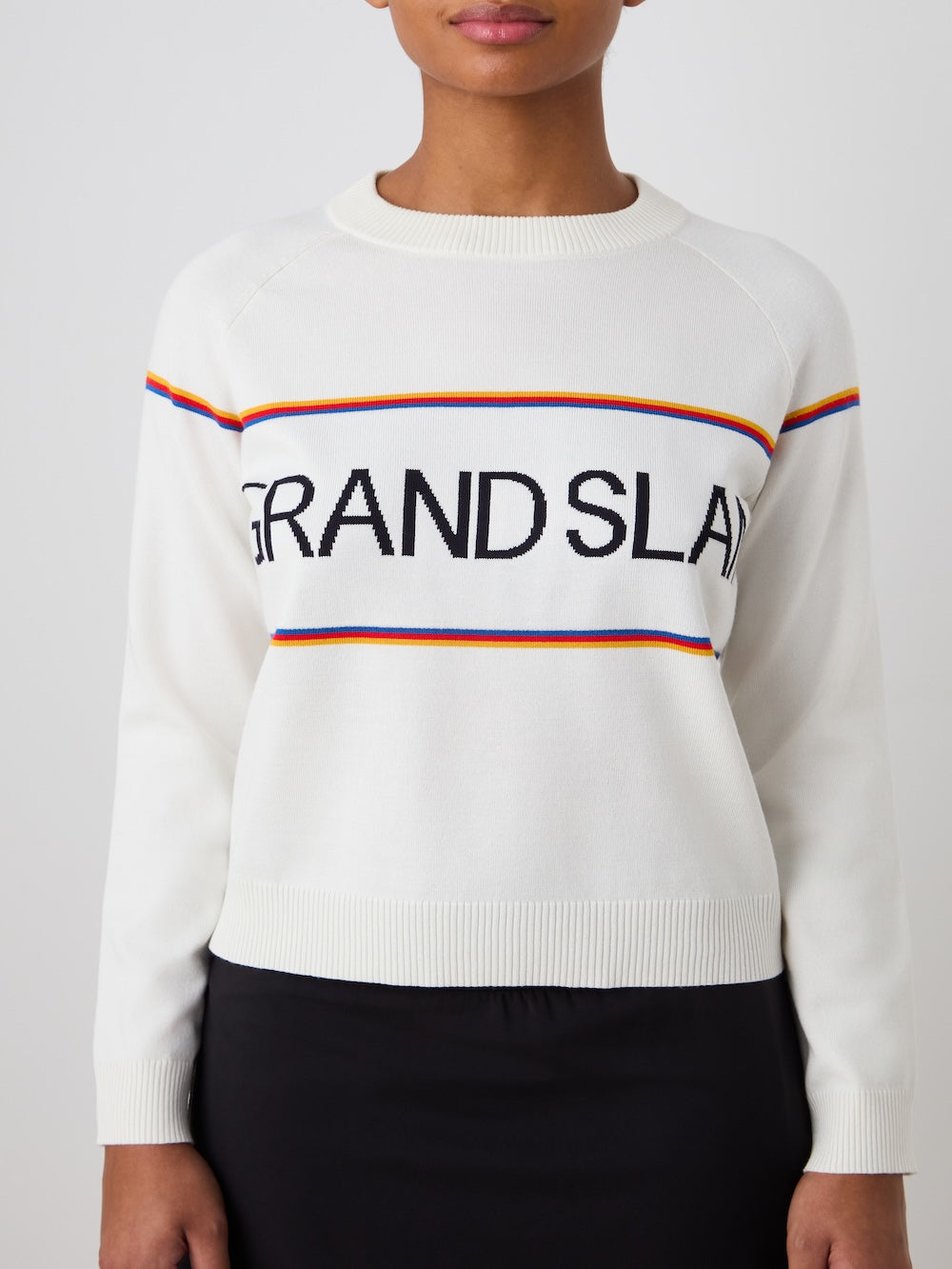 A person wears the We Norwegians Grand Slam Sweater Women—a white, vintage athletic après sport layer with "GRAND SLAM" in black and thin red, blue, and yellow stripes—paired with a black skirt.