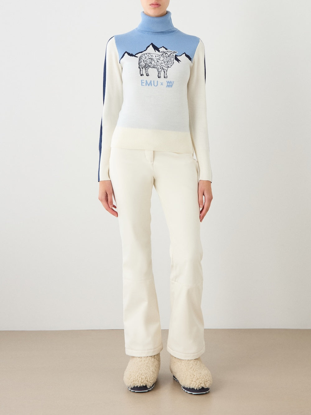 A person wearing the EMU x WN Ski Sweater Women by We Norwegians, featuring a charming sheep and mountain design in blue and white, paired with white pants and fluffy beige boots from the EMU x WN collaboration, stands on a tan floor against a plain white wall.