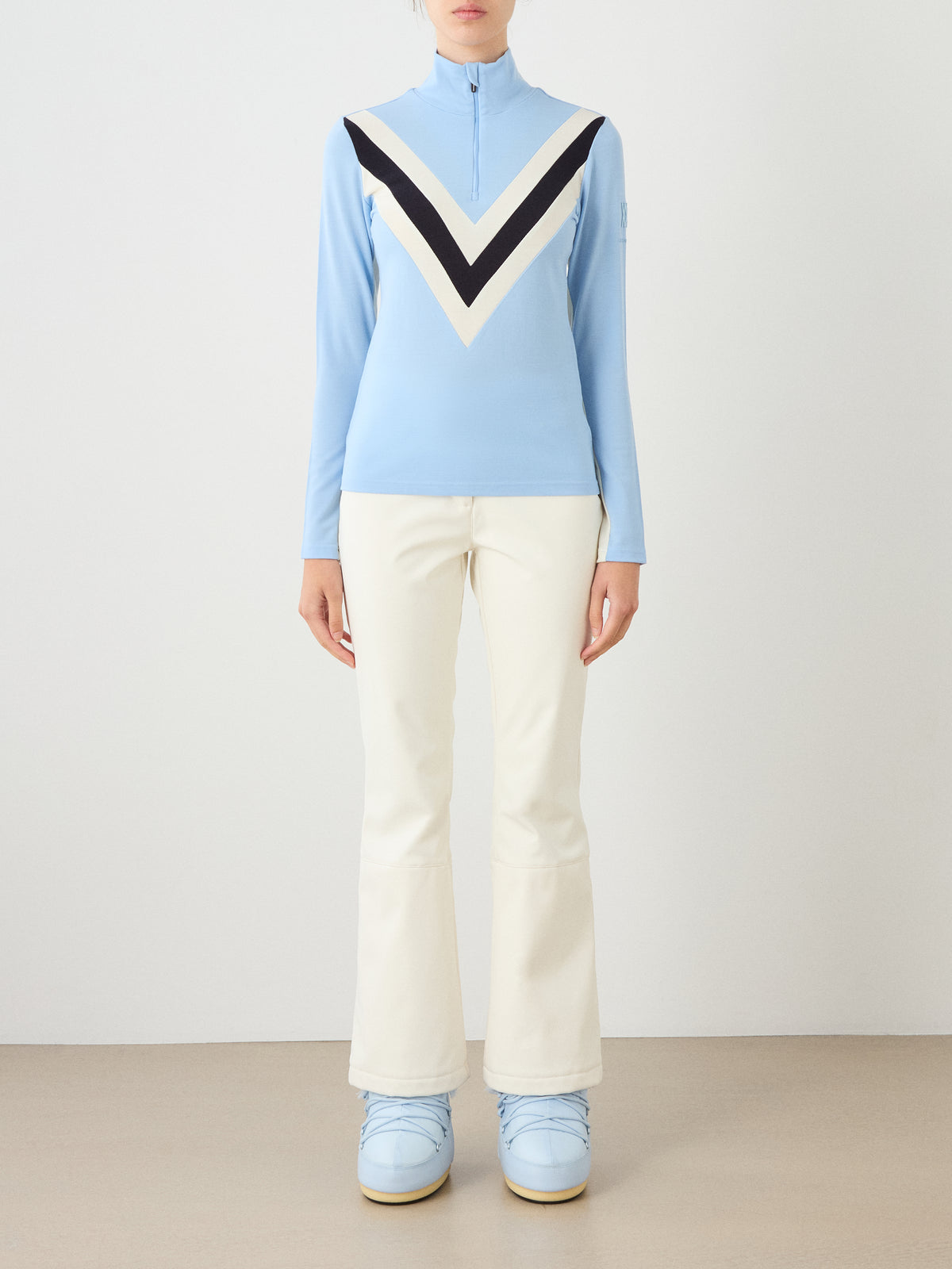 A person wears a light blue We Norwegians Voss Chevron ZipUp Women zip-up with a black and white chevron pattern on the chest, paired with off-white flared trousers and light blue boots.