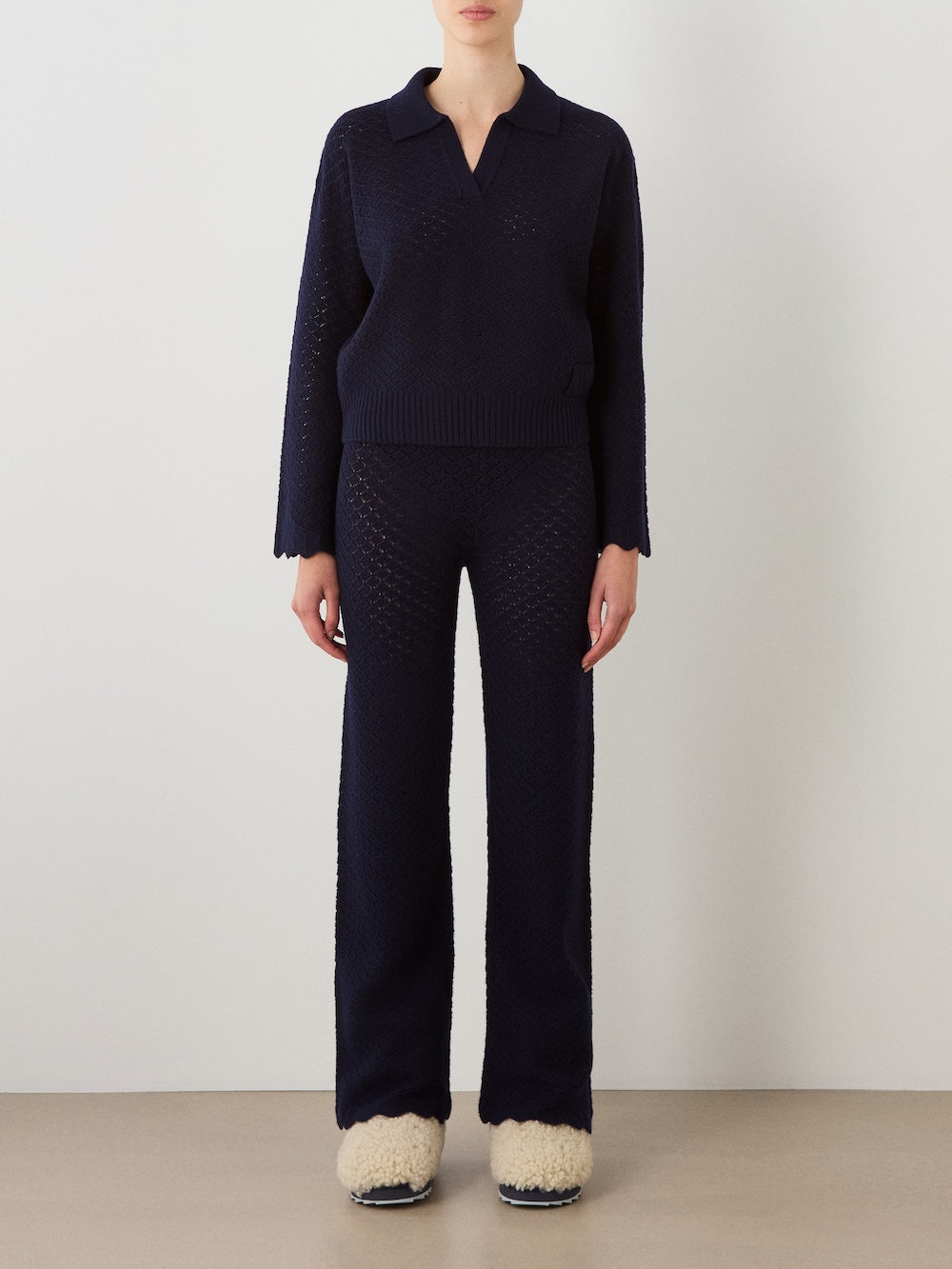 A person stands against a plain wall wearing the Saeter Henley Women sweater by We Norwegians, paired with matching knit pants and cream-colored fuzzy slippers. Their hands rest by their sides.