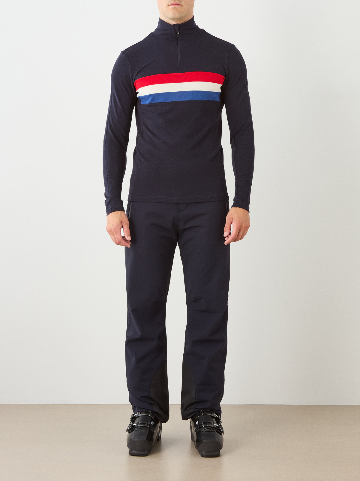 A person wearing black ski boots, navy pants, and the We Norwegians Ski ZipUp Men—a navy long-sleeve top featuring red, white, and blue horizontal stripes across the chest—stands against a plain white wall, evoking classic retro-inspired ski wear.