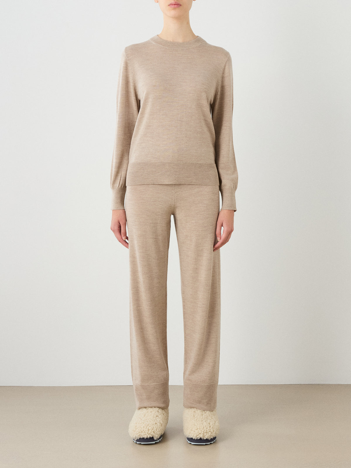A person stands against a plain background wearing the We Norwegians Geilo Crewneck Women sweater in beige, paired with matching beige pants and cream-colored fuzzy slippers. Their hands hang relaxed at their sides, and their face is not visible.