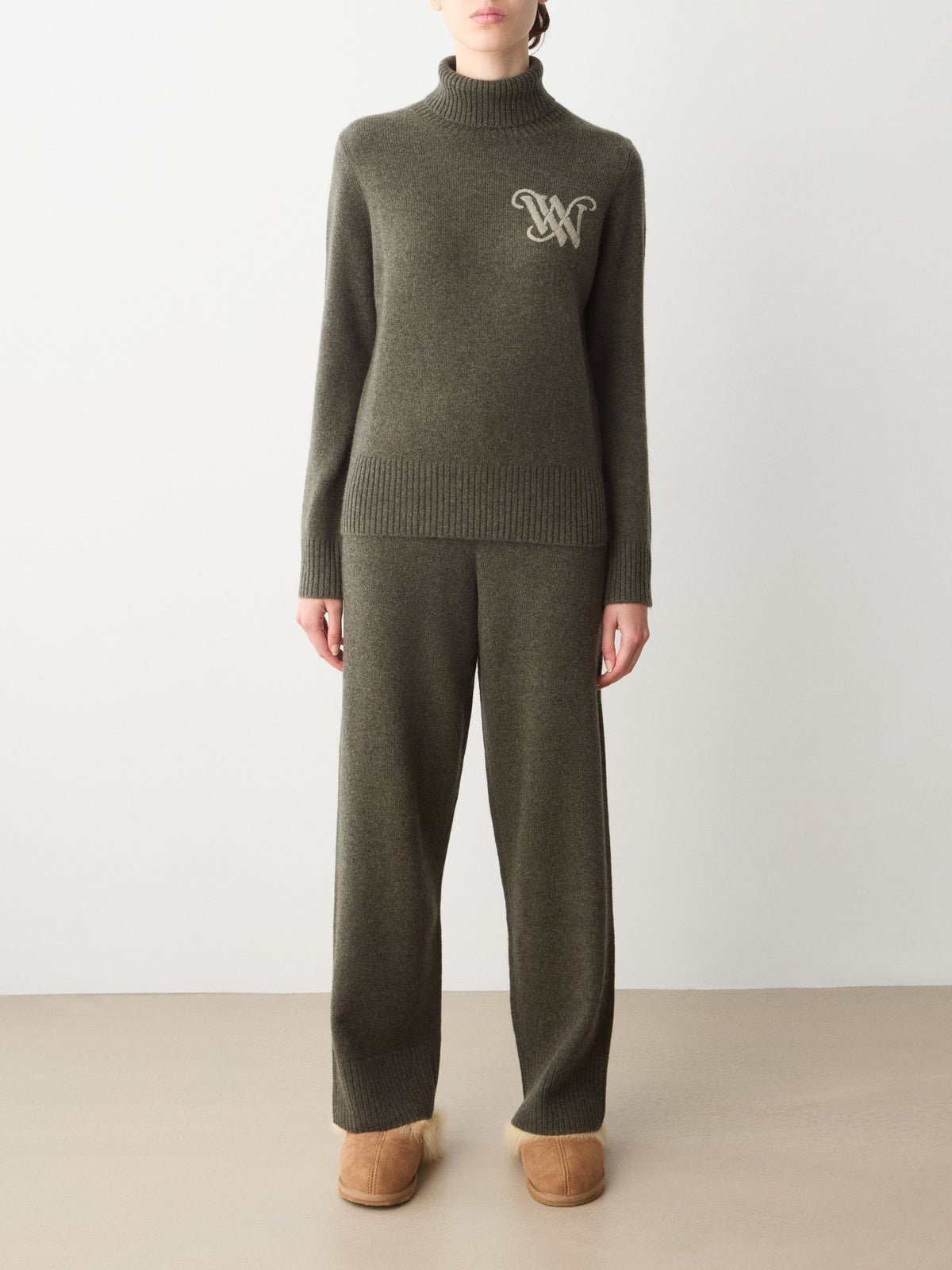 A person wearing a dark olive We Norwegians Monogram Cashmere Sweater Women, matching wide-leg trousers, and tan slippers.