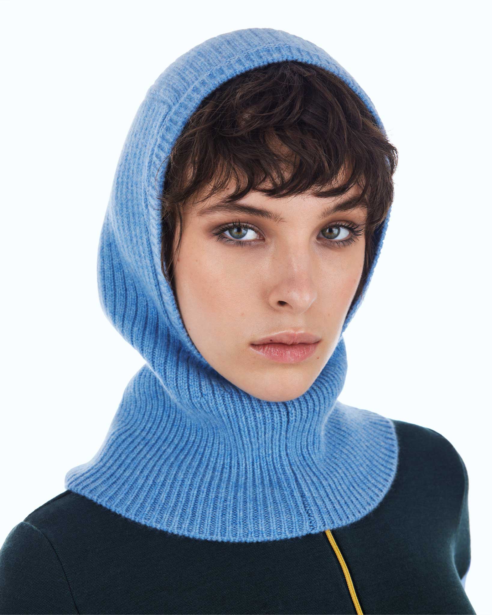 A person with short, dark hair is wearing a light blue Blefjell Balaclava Women by We Norwegians, stylishly knitted in a cozy 1/2 cardigan stitch. They are dressed in a dark top featuring a yellow zipper. The background is plain and light-colored, which draws attention to the person's face and the distinctive balaclava.