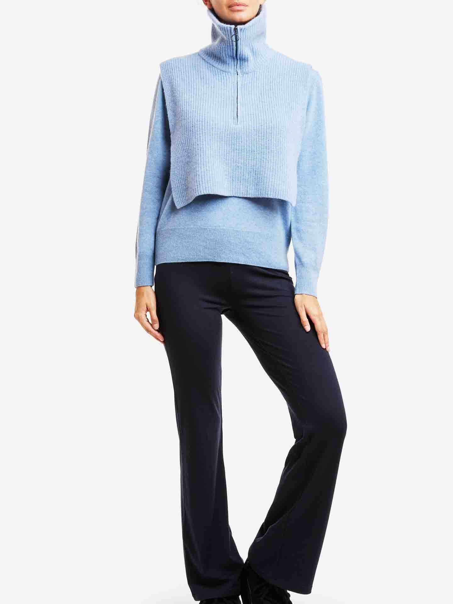 A person wearing the Blefjell Cashmere Bib Women by We Norwegians in light blue, paired with a layered vest and navy flared pants, stands against a plain background.