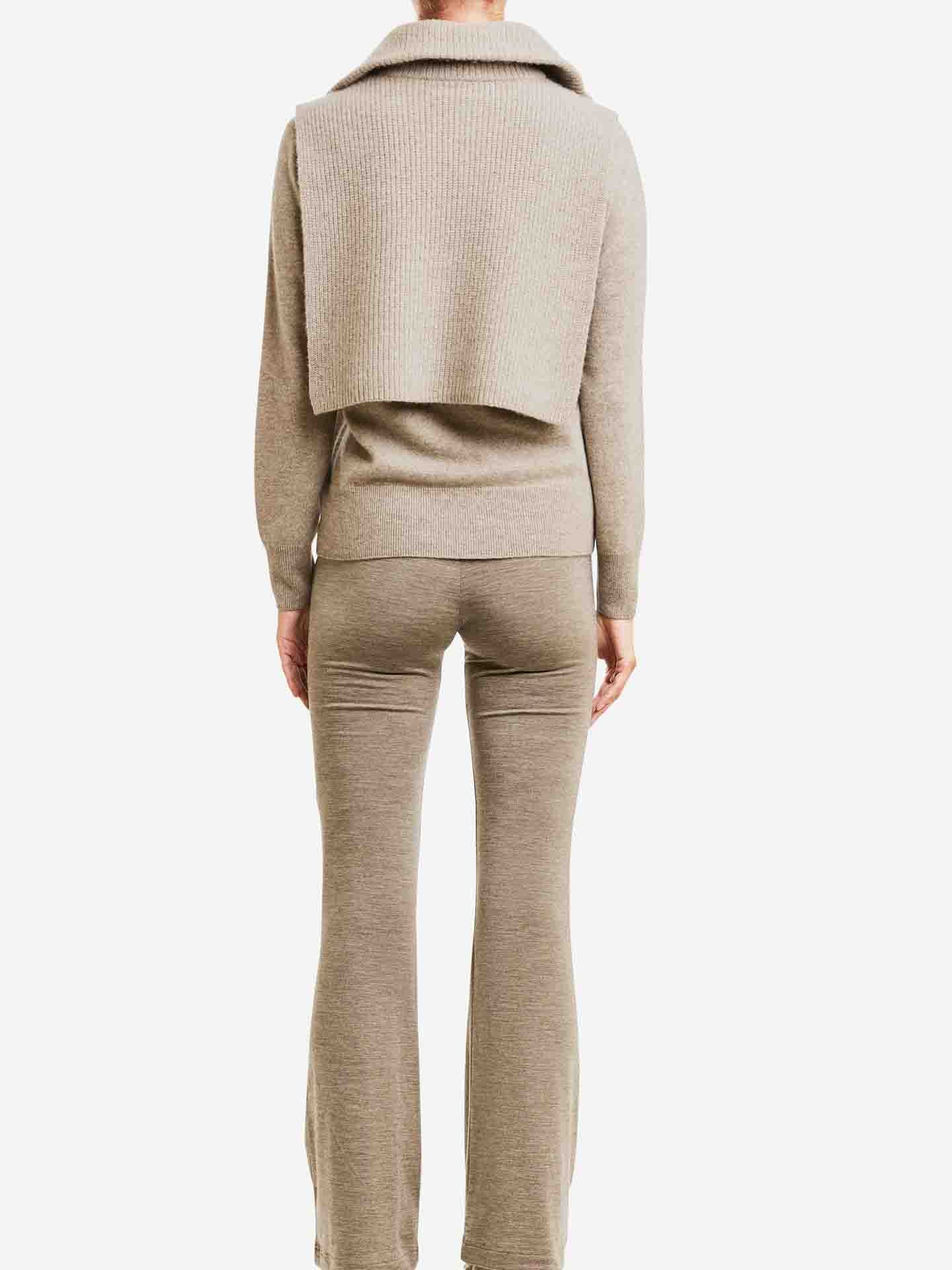 A person is seen from the back wearing the We Norwegians Blefjell Cashmere Bib, a beige knit sweater featuring matching wrap details and flared pants, all topped with a layered vest, set against a plain white background.