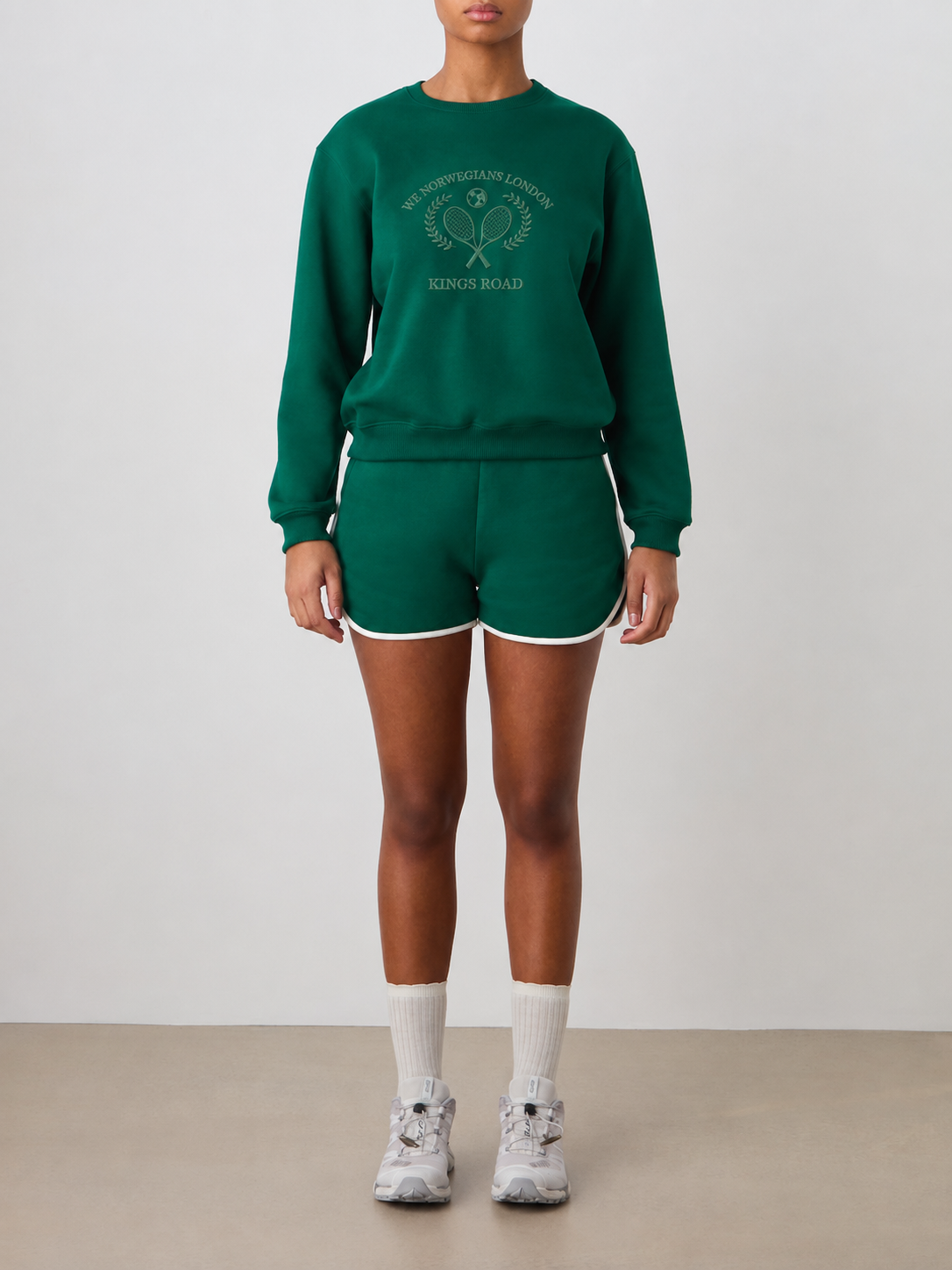 Wearing the We Norwegians Chelsea Sweater Women in a relaxed fit green cotton crew neck with tennis embroidery and matching shorts, a person stands against a plain background with hands at their sides, paired with white socks and sneakers.
