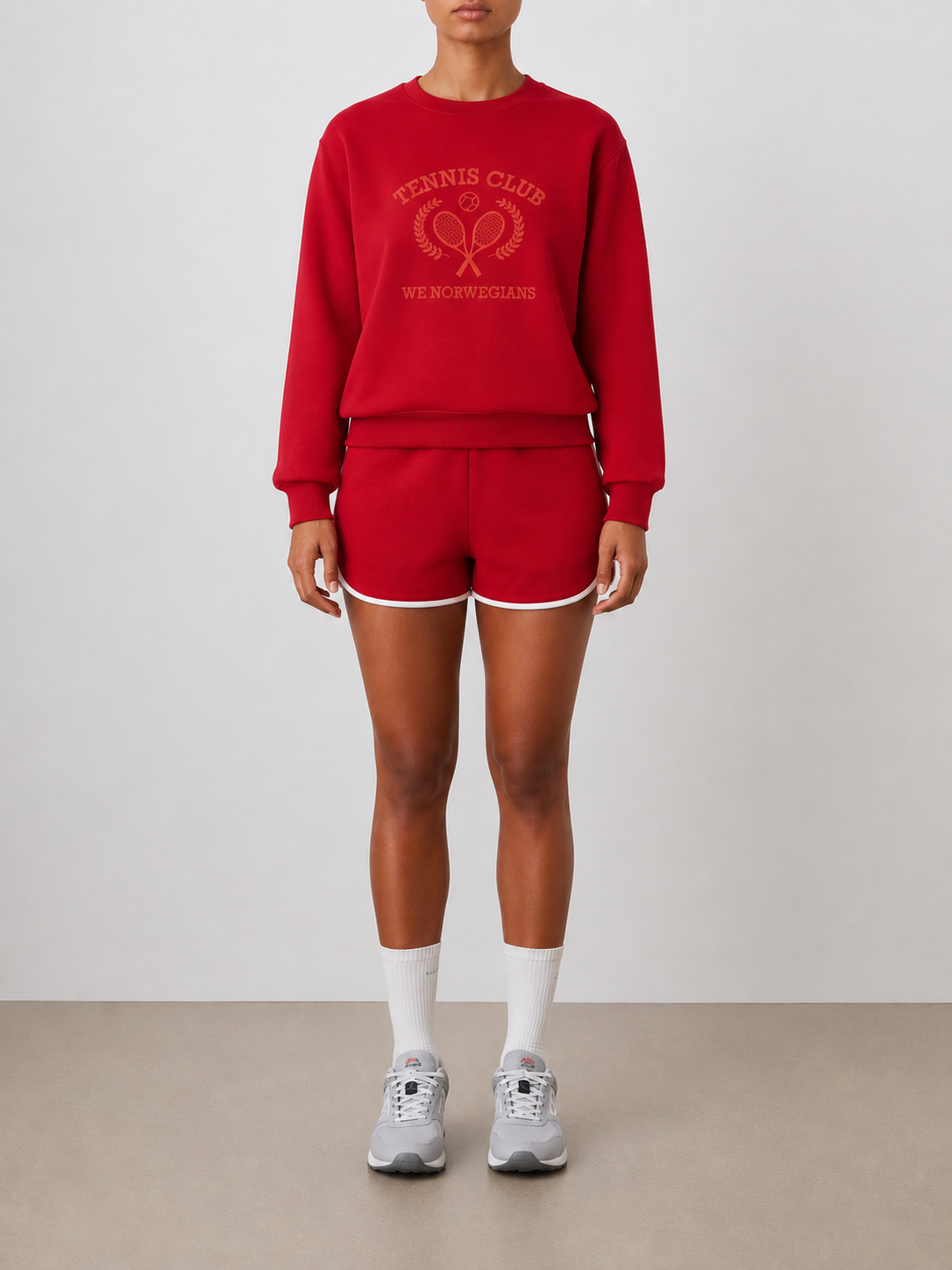 A person stands against a plain background wearing the Chelsea Sweater Women by We Norwegians, a red cotton jersey crew neck sweatshirt with "Tennis Club WE NORWEGIANS" and tennis embroidery, paired with matching red shorts, white socks, and gray sneakers.