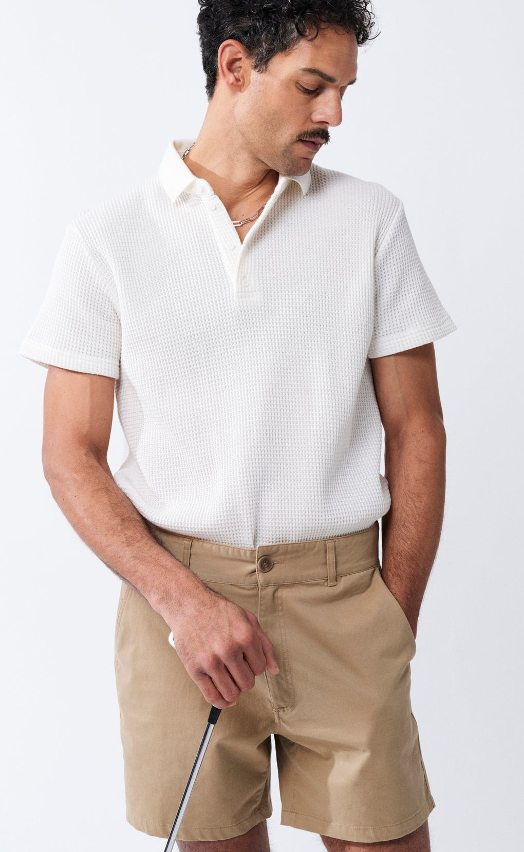 Man wearing a white polo shirt and beige shorts holding a golf club on a white background