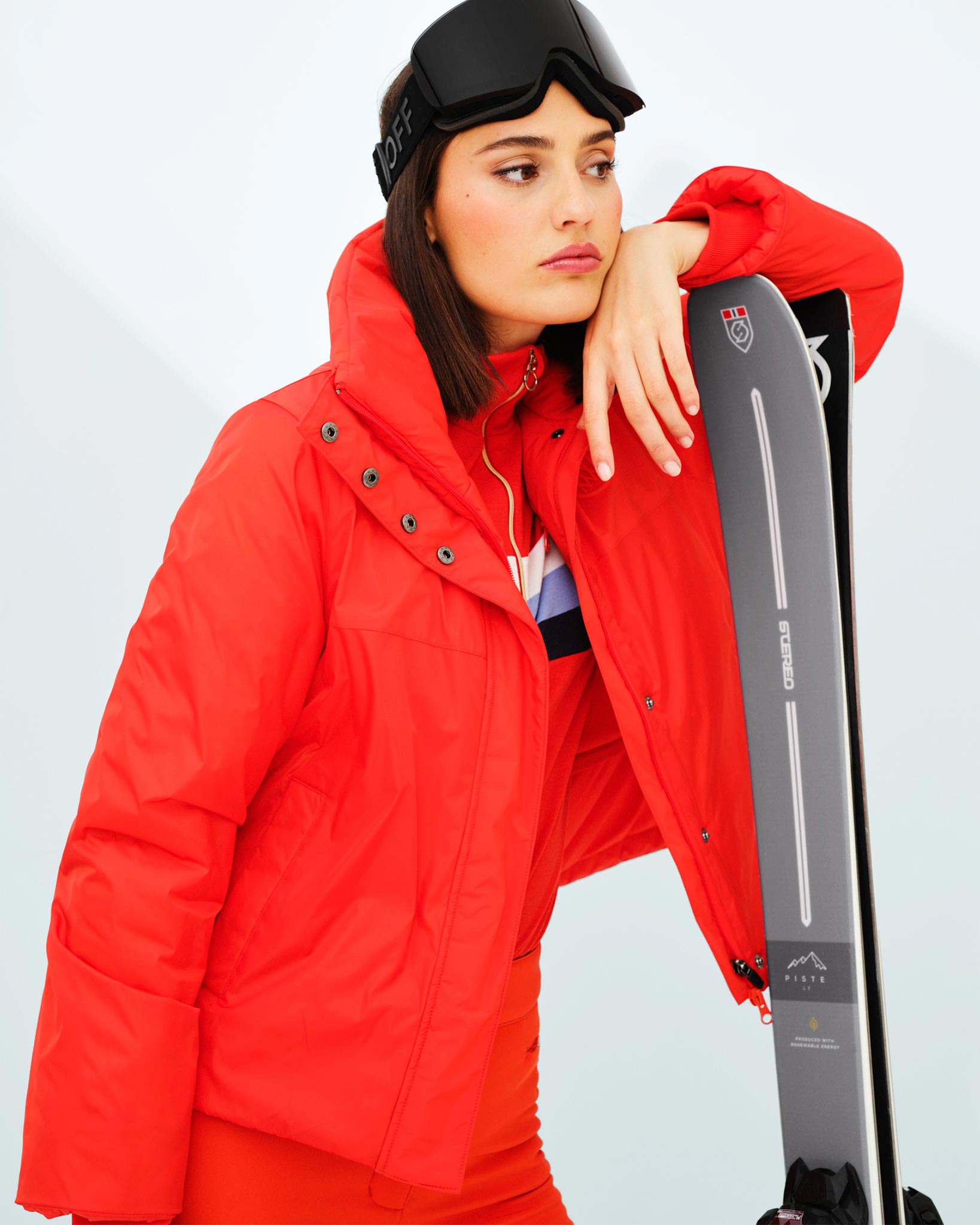 A woman in a bright red We Norwegians Filefjell Ski Jacket and pants, made with waterproof fabric and wool padding, is leaning on a pair of skis, looking contemplative. She is wearing black ski goggles on her head, and her dark hair is down. The background is a minimal, light-colored indoor setting.