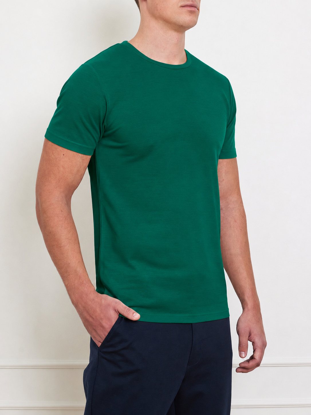 A man wears the We Norwegians Medalist Piqué Tee Men, a plain green moisture-wicking golf T-shirt, with dark pants. He stands against a light wall, one hand in his pocket, his face partially out of frame.