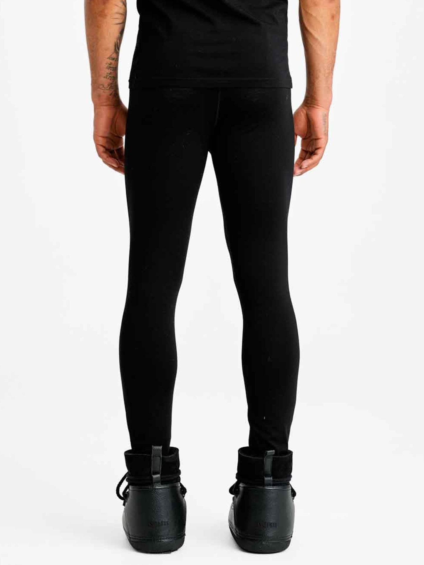 The image shows the legs and lower back of a person wearing We Norwegians Sno Merino Long Johns Bottoms Men, made from breathable fabric, along with dark green high-top sneakers. The person is standing facing away from the camera against a plain white background with visible tattoos on their arms.