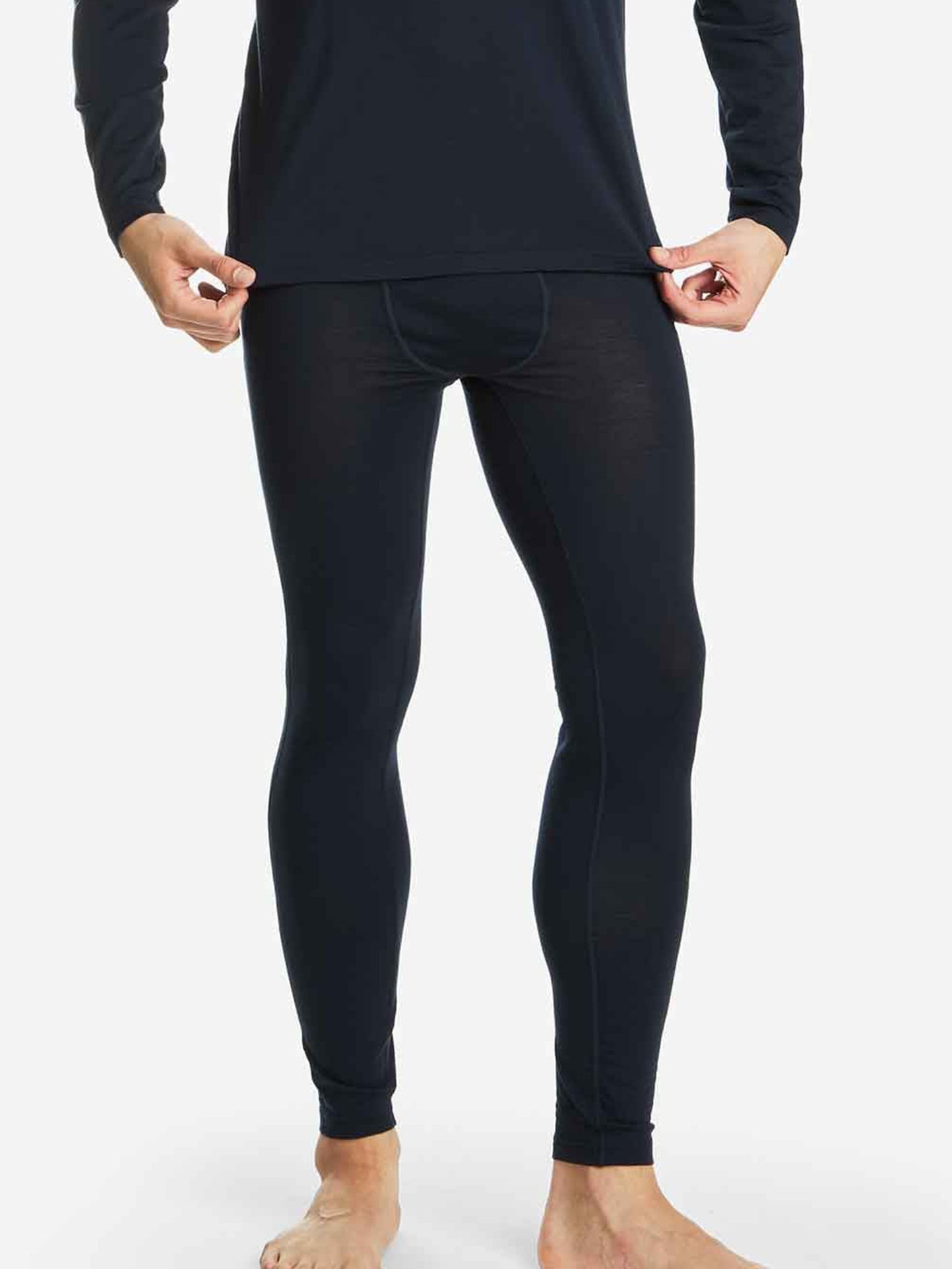 A person is wearing a black long-sleeve top and black Sno Merino Long Johns Bottoms for men by We Norwegians. The person is barefoot and is holding the hem of the top slightly raised with both hands. The background is plain white.