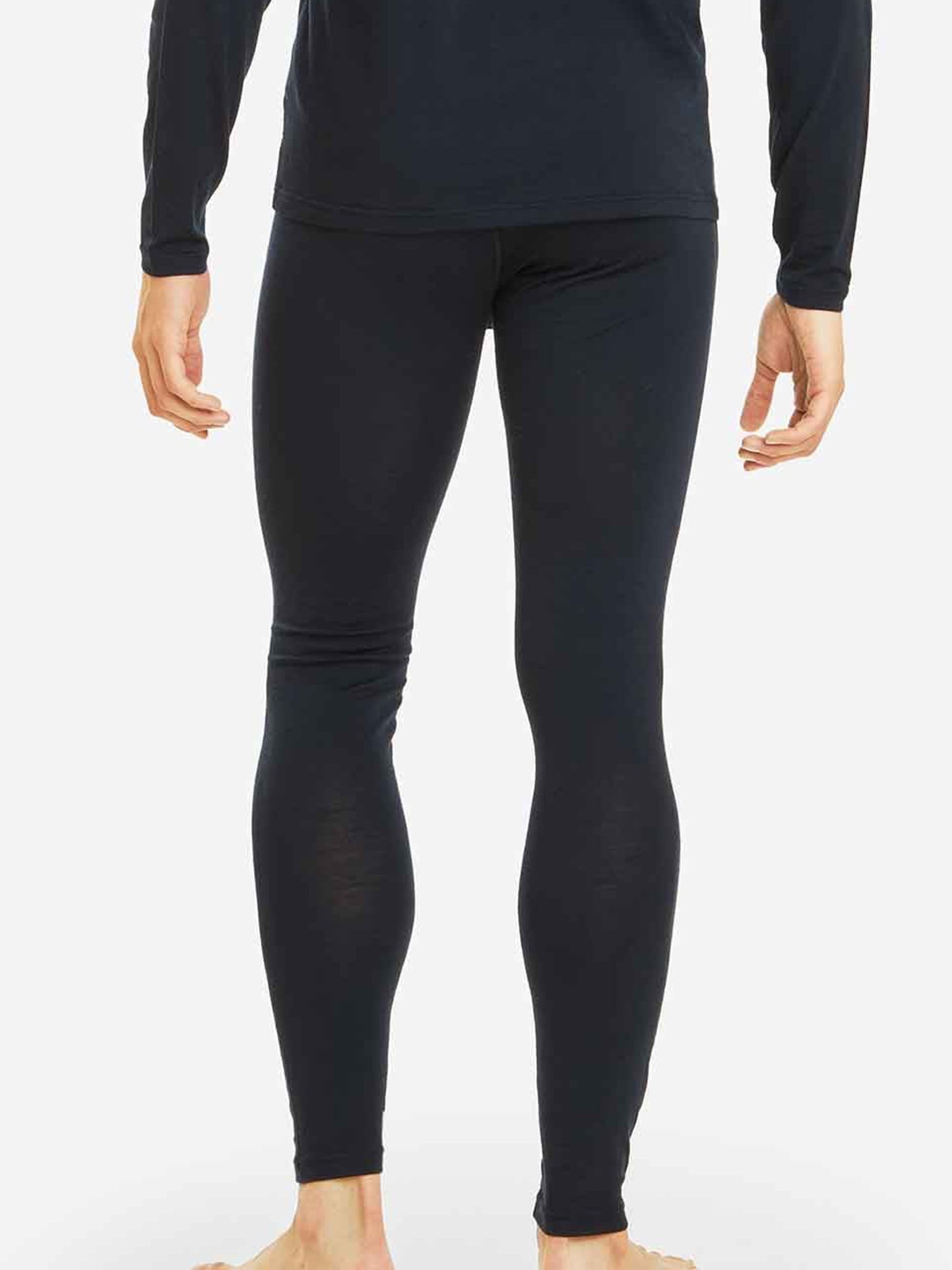 A person clad in We Norwegians' Sno Merino Long Johns Bottoms Men and a long-sleeve black top, standing barefoot on a light-colored floor. The image is captured from the back, highlighting the full length of the breathable base-layer leggings and part of the top.