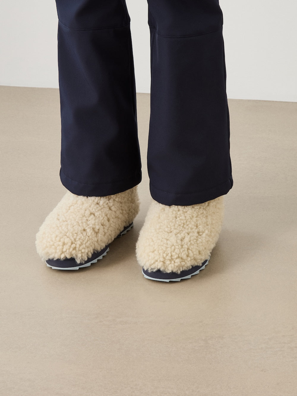 A person wearing dark pants and cream-colored, fluffy EMU x WN Sharky Lo Teddy slippers by We Norwegians—crafted from luxurious Australian sheepskin with black soles—stands on a beige floor.