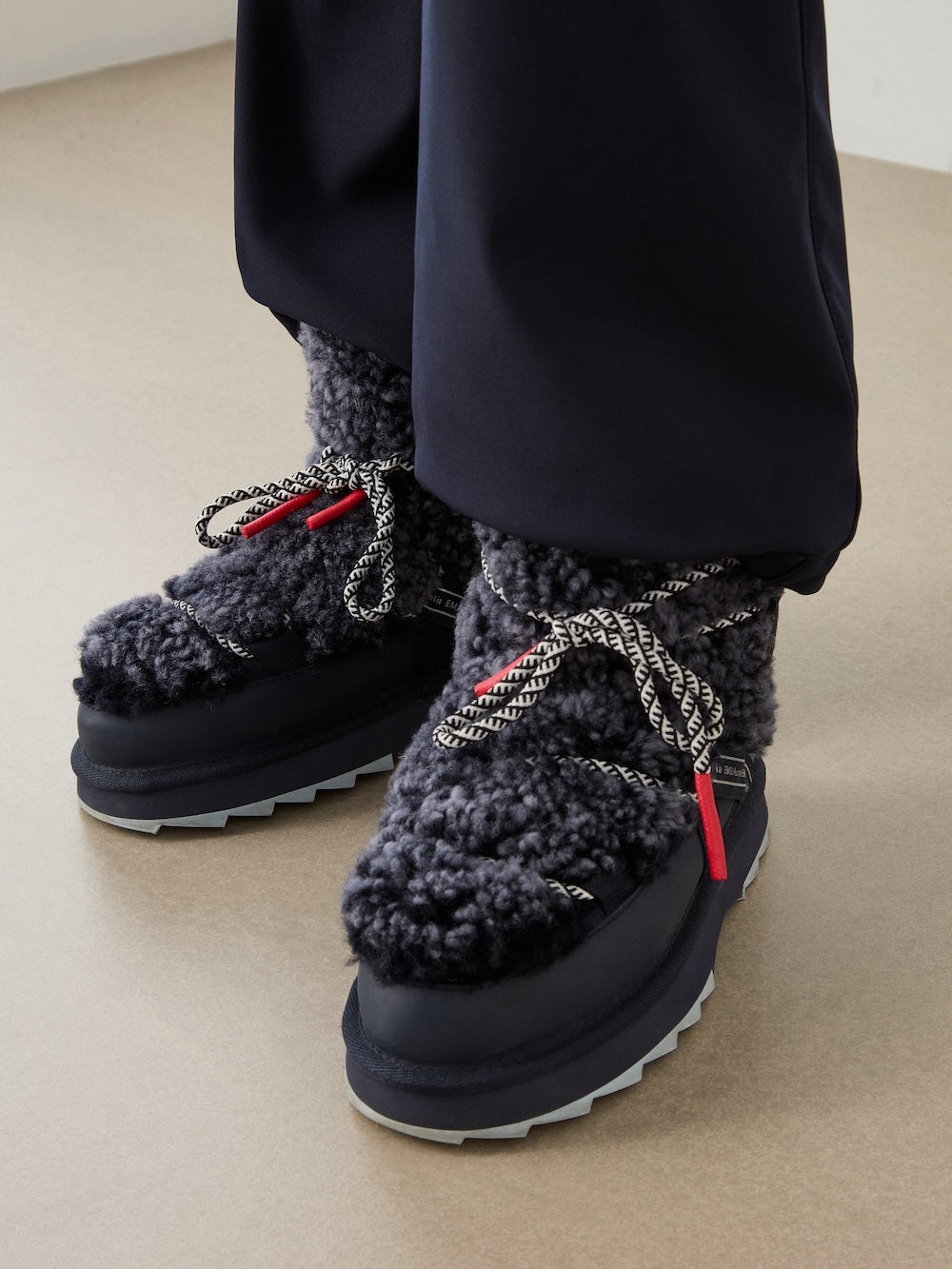A person wearing EMU x WN Blurred Teddy sheepskin boots by EMU, featuring a dark, fluffy plush exterior with thick patterned laces and red lace tips, paired with black pants on a beige floor.