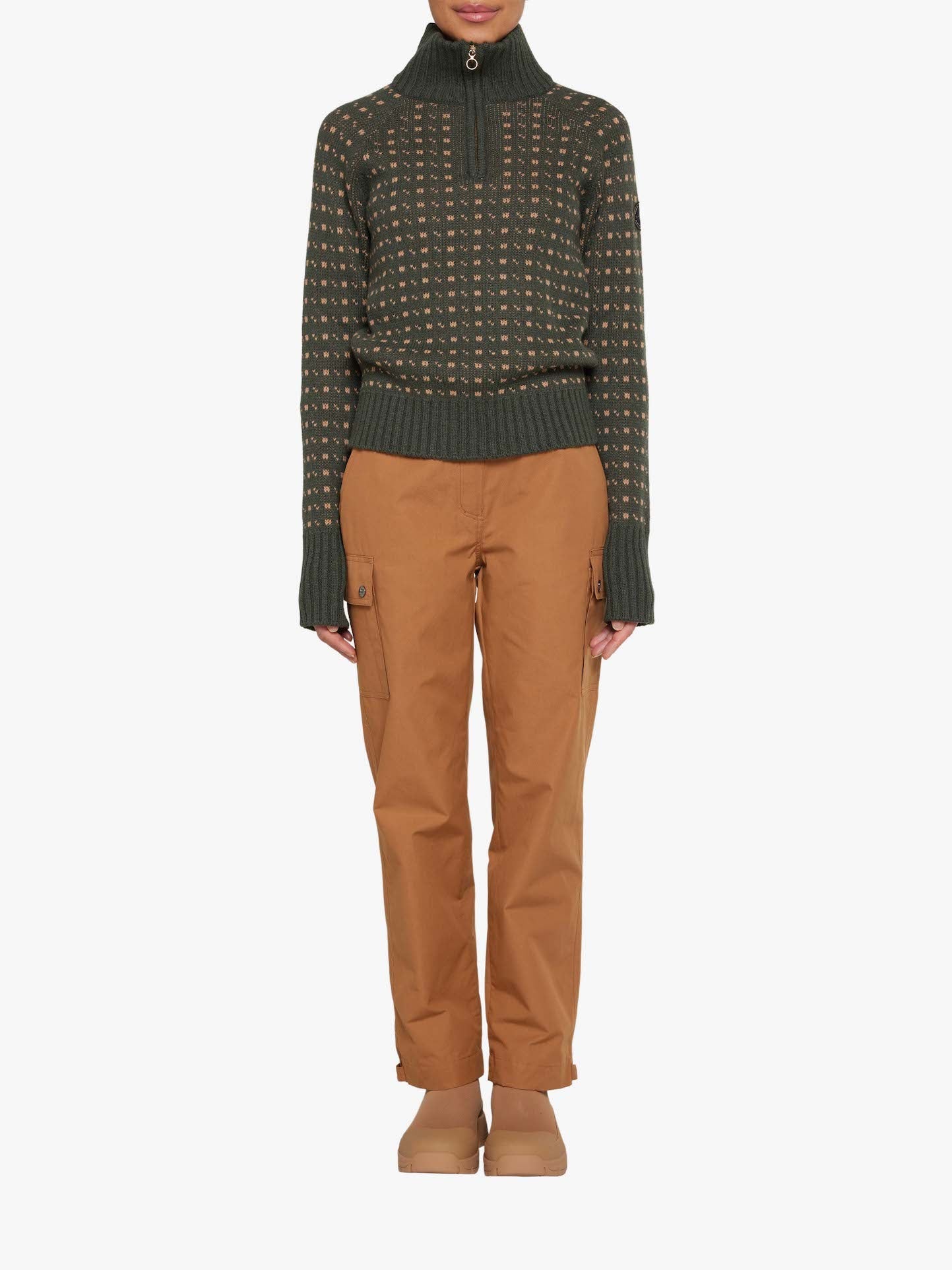 A person is standing against a plain white background, wearing a dark green turtleneck sweater with a small pattern, made of Ventile® fabric prized for its weatherproof qualities. They are also dressed in We Norwegians' Lyngen Pant Women in Cinnamon and light brown shoes. The sweater features a half-zip design at the neck, reminiscent of those worn by RAF pilots. Their hands are by their sides.