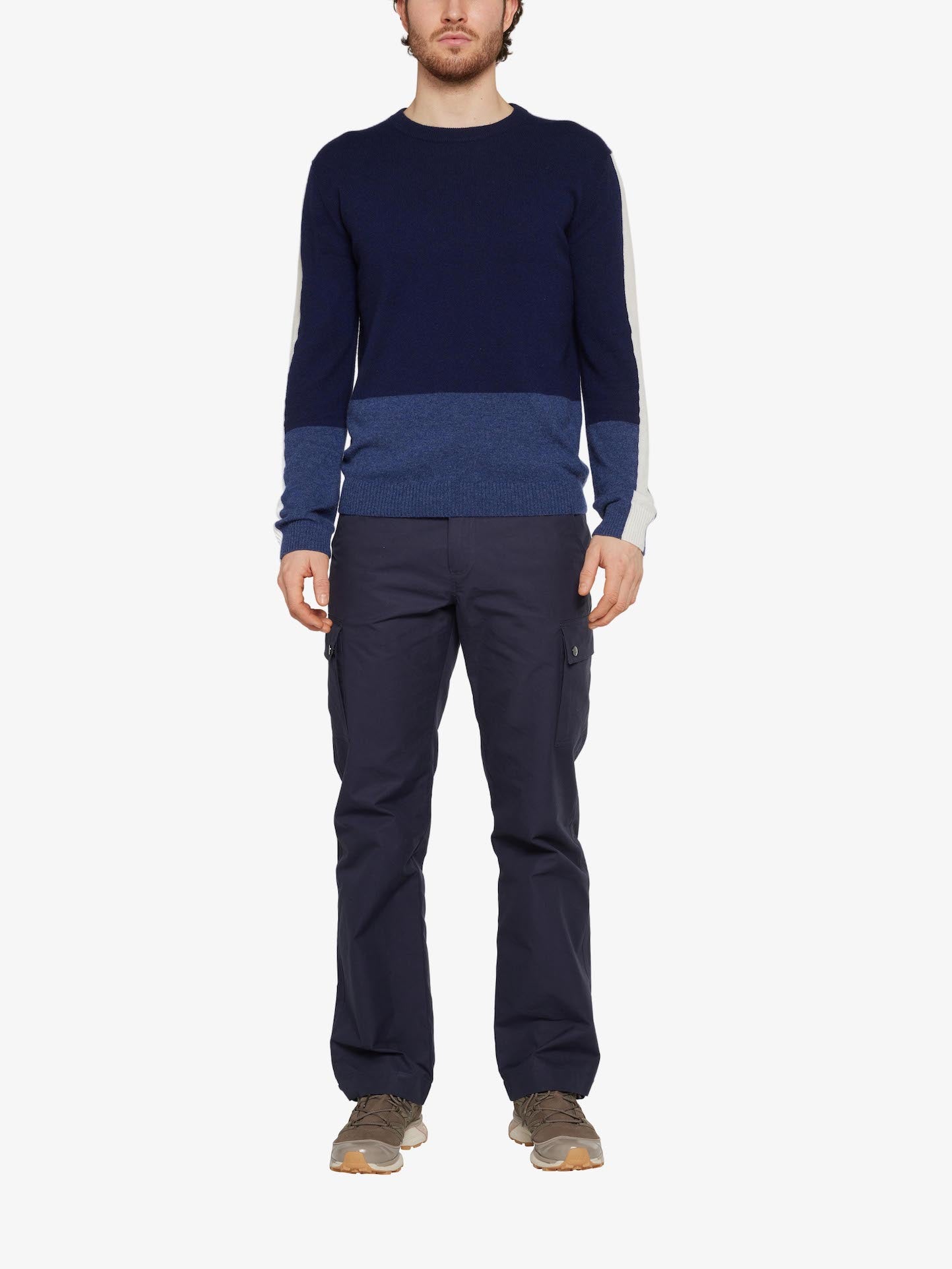 A man is standing against a plain white background wearing a color-block sweater in shades of navy blue, royal blue, and white. He's also wearing We Norwegians' Lyngen Pant Men Navy Blue cargo pants made from Ventile® cotton fabric known for its weatherproof qualities and light brown shoes. His hands are at his sides, and he is facing forward.