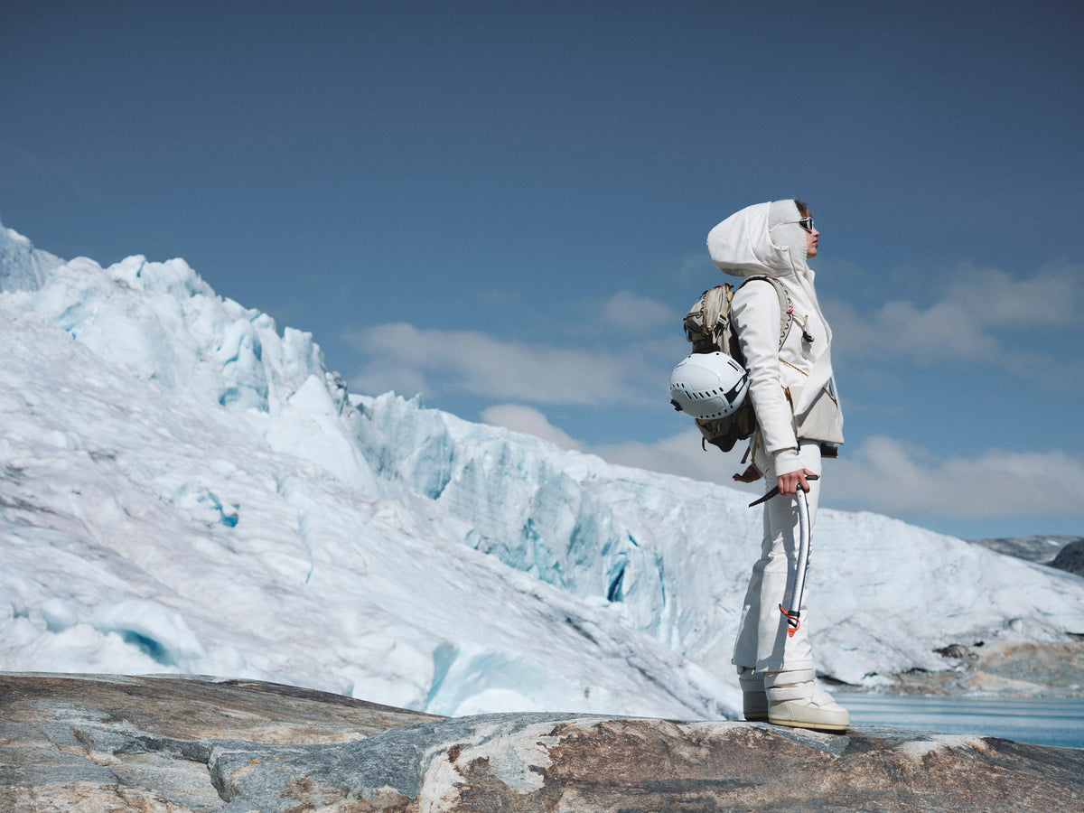 A person in white winter gear stands on a rocky surface with a backpack and helmet, gazing at a snowy glacier under a clear blue sky.