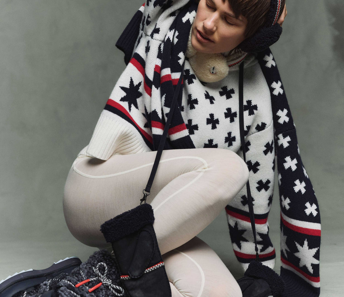 A person in earmuffs, a patterned sweater with stars and crosses, cream leggings, and winter boots kneels with one arm raised, eyes closed, against a neutral backdrop.
