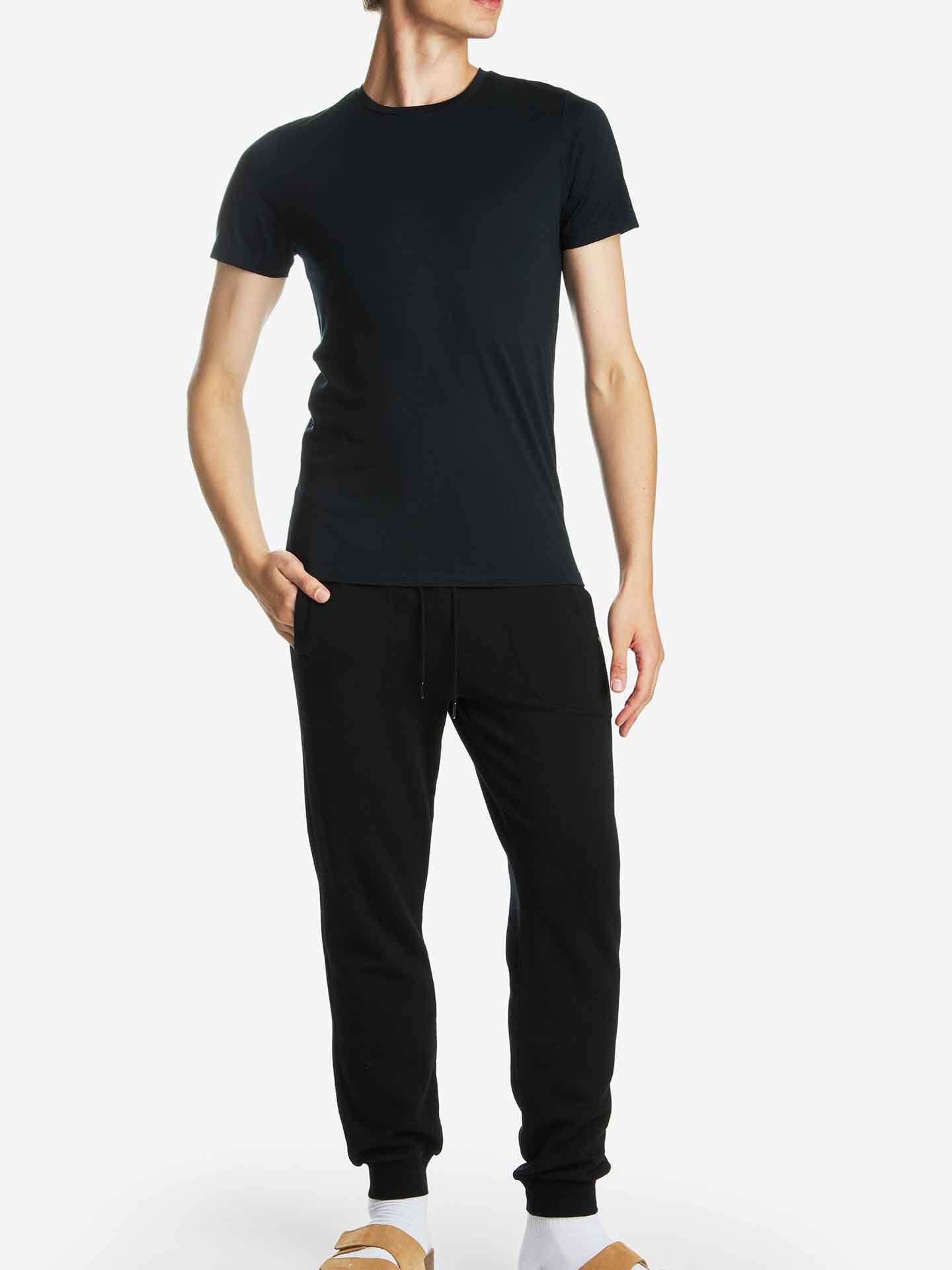 A person is standing against a plain white background wearing a We Norwegians Base One Tee Men, black jogger pants, and tan slippers with white socks. They are posing with one hand in their pocket and the other resting by their side. Their face is not visible in the image.