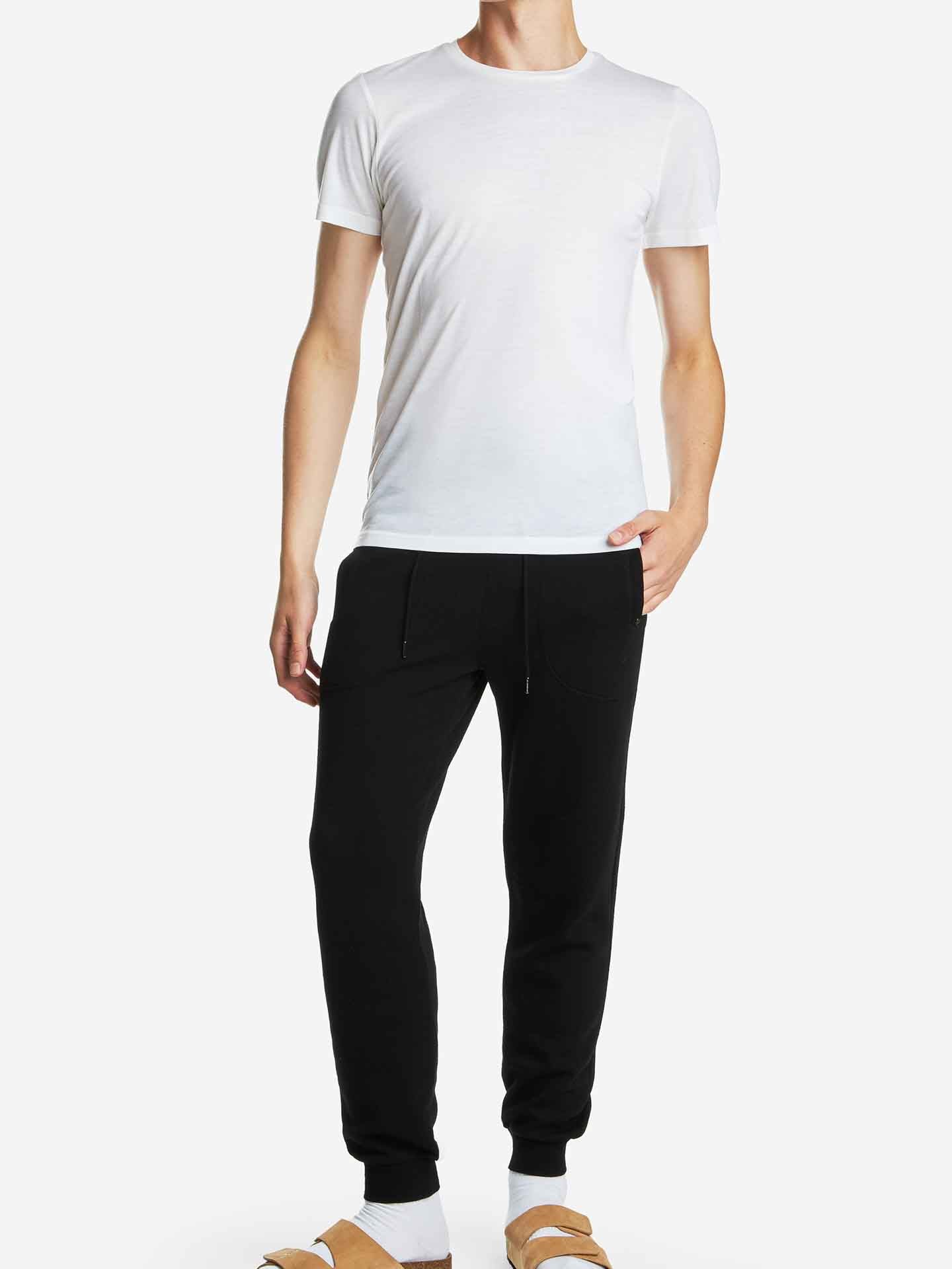 A person is standing wearing a plain white short-sleeve Base One Tee from We Norwegians and black jogger pants made from 100% merino wool. They have one hand in their pocket. They are also wearing white socks and brown slide sandals. The background is plain white. The person's head is not visible in the image.