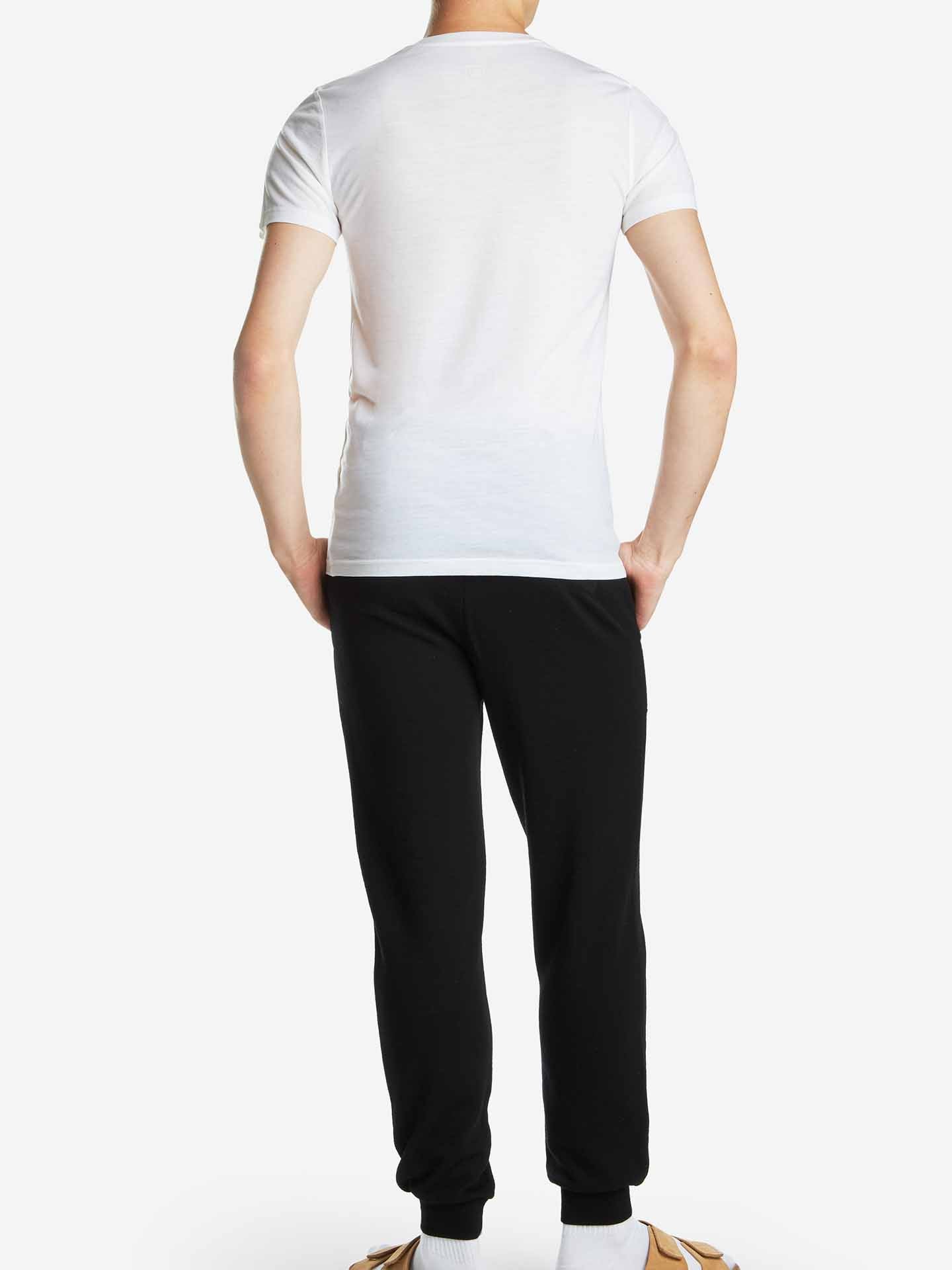 A person is standing with their back to the camera, wearing a We Norwegians Base One Tee Men, which is a white short-sleeved, odor-resistant t-shirt made of 100% merino wool. They are also wearing black pants and tan shoes against a plain white background.