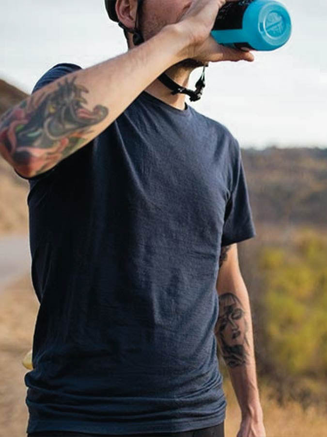 A person wearing a dark blue Base One Tee Men by We Norwegians and a bicycle helmet drinks from a blue water bottle. The individual, adorned with several arm tattoos, is situated in an outdoor environment that appears to be a hiking or biking trail with dry grass and gentle hills.