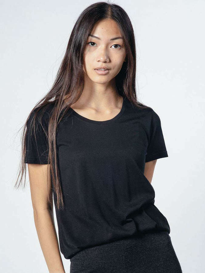 A person with long, straight hair is standing against a plain white background. They are wearing a black short-sleeve Base One Tee Women by We Norwegians and dark pants. Their expression is neutral, and their hands are resting by their sides.