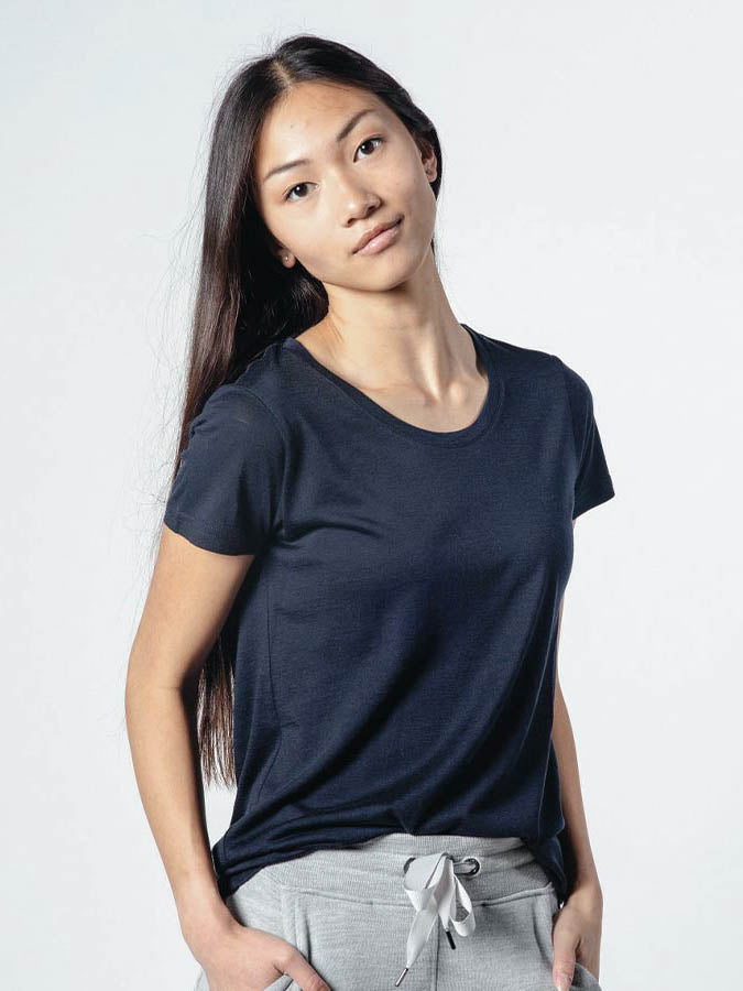 A person with long, straight hair stands against a plain background. They are wearing the dark, luxurious We Norwegians Base One Tee Women and light-colored pants with a bow at the waistband. The individual has a neutral expression and their hands are in their pockets.