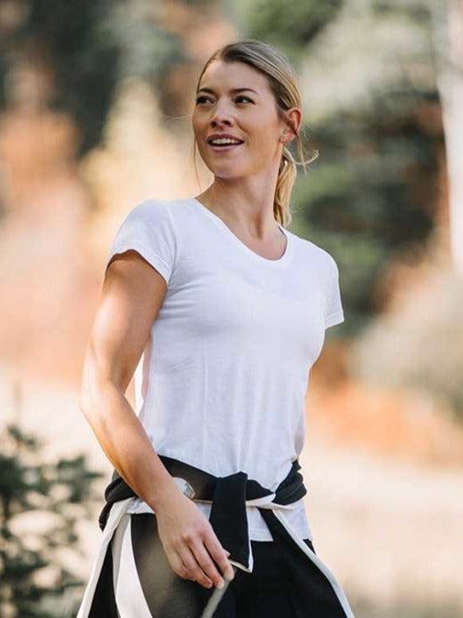 A woman with blonde hair tied back is walking outdoors with a light smile, wearing the luxurious We Norwegians Base One Tee Women in white and a black jacket tied around her waist. The background is blurred with natural, autumnal scenery.