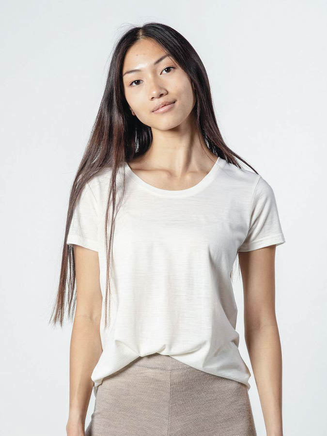 A person with long, straight hair, wearing the Base One Tee Women by We Norwegians and beige pants stands against a light gray background, looking towards the camera with a neutral expression.