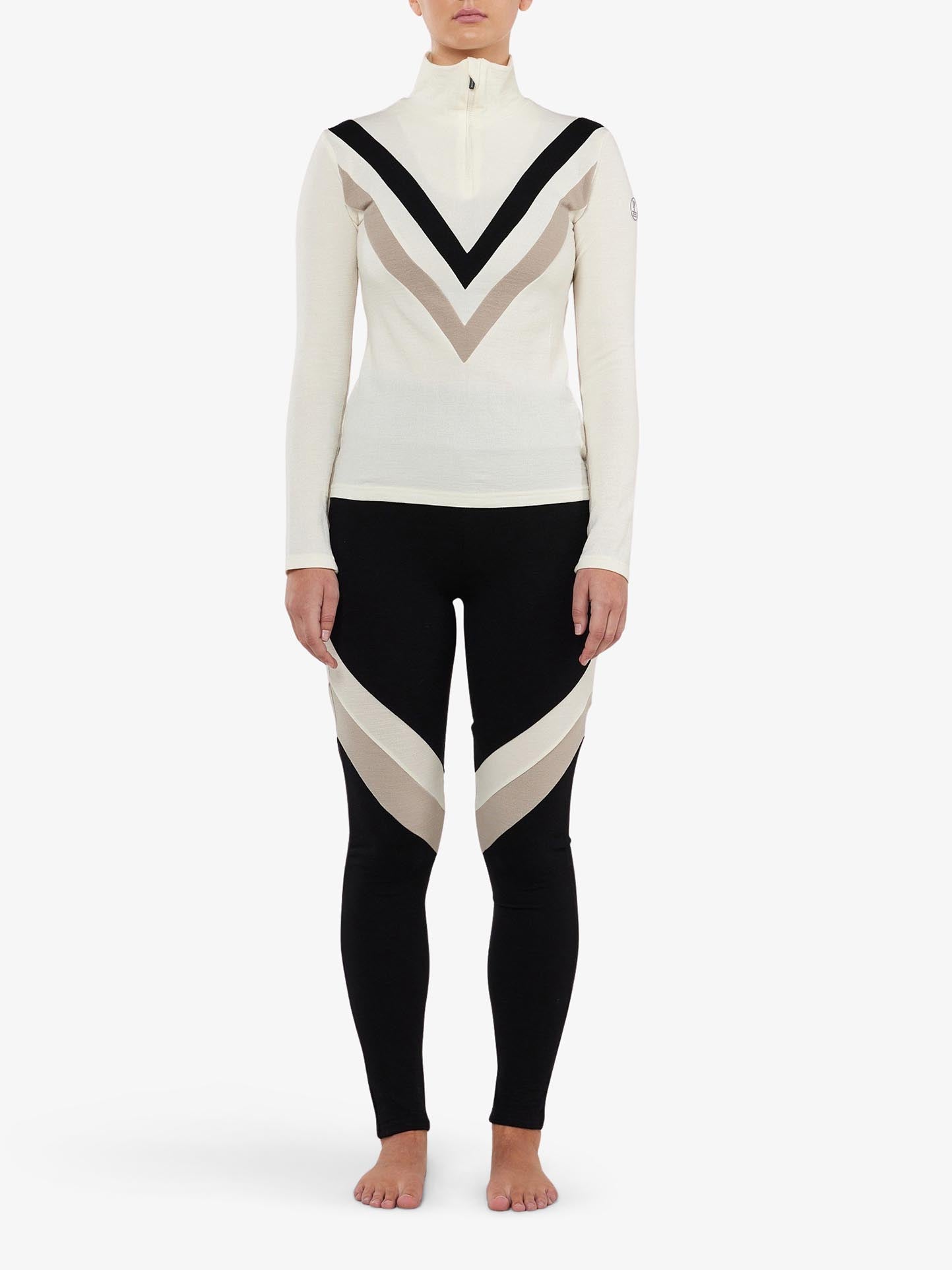 A person stands against a white background wearing a long-sleeve, high-neck, zippered top with black and beige V-shaped patterns. They are also sporting the Voss ColBlock Leggings Women by We Norwegians, featuring beige and white V-shaped designs. The merino wool base-layer ensemble is stylish and functional, highlighting their barefoot elegance.