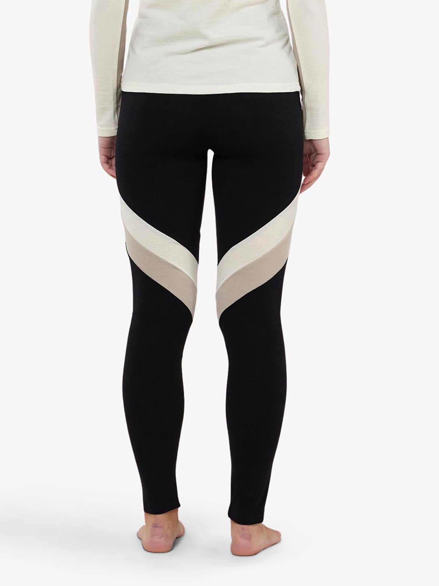 A woman is wearing We Norwegians Voss ColBlock Leggings Women featuring beige diagonal stripes on the thighs. She is barefoot and standing with her back facing the camera, highlighting a long-sleeve off-white top ideal for base-layer skiing. The background is white.