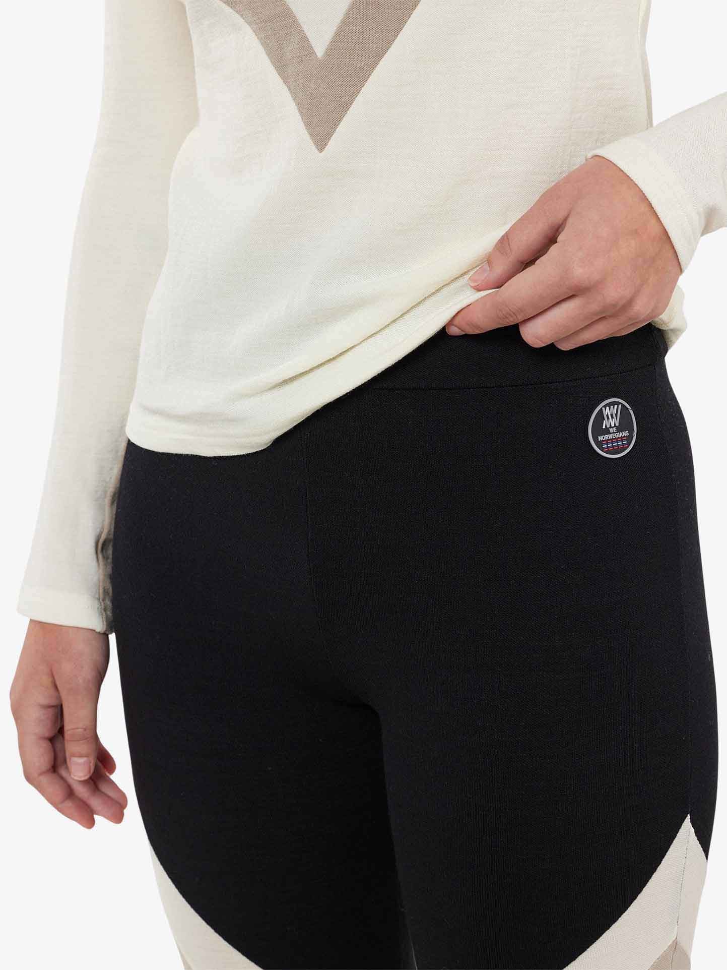 A person wearing a white long-sleeve shirt and We Norwegians Voss ColBlock Leggings Women, featuring beige accents. The hand is holding the hem of the shirt, and a small circular logo is visible on the waistband of these merino wool leggings, perfect for base-layer skiing.