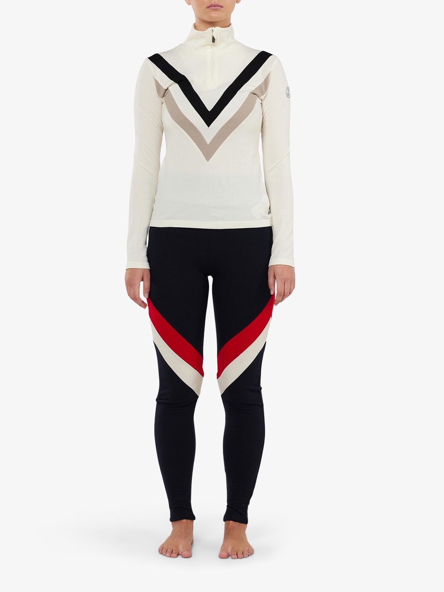 A person is standing barefoot against a white background, wearing a long-sleeve, beige merino wool turtleneck top with a distinctive V-shaped design in black and white on the chest. They are also wearing We Norwegians' Voss ColBlock Leggings Women, featuring diagonal red and beige stripes on the thighs.