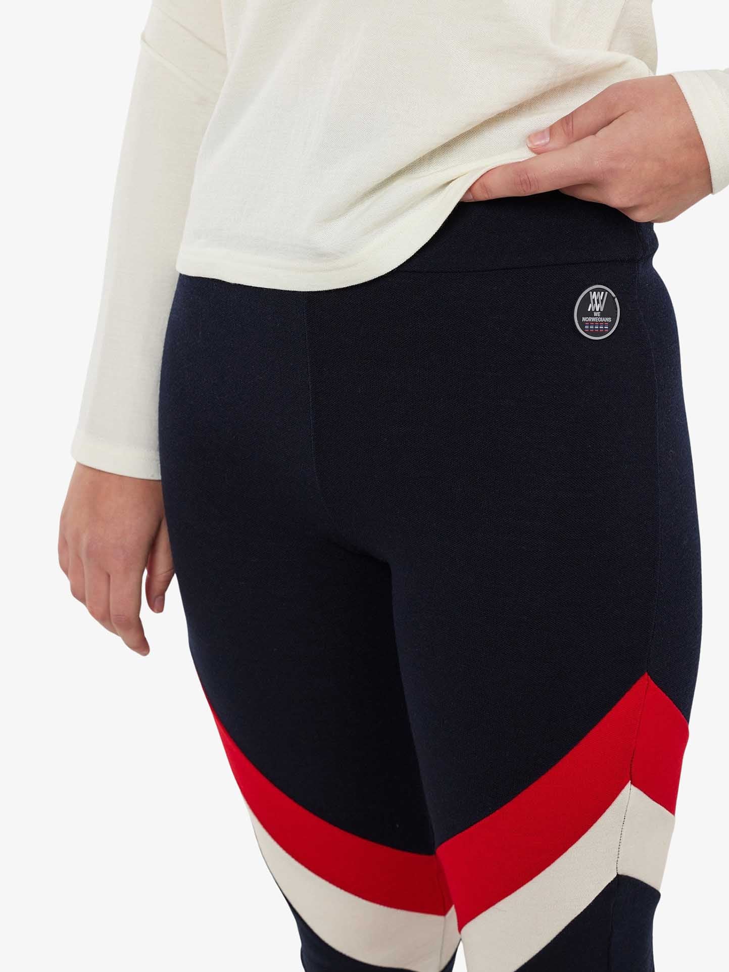 A person is wearing Voss ColBlock Leggings Women in navy blue with red and white diagonal stripes near the knees by We Norwegians. Their left hand is resting on their hip, complementing a white long-sleeve top. The leggings feature the brand's logo with two overlapping W's, making it an ideal stylish base-layer for skiing.