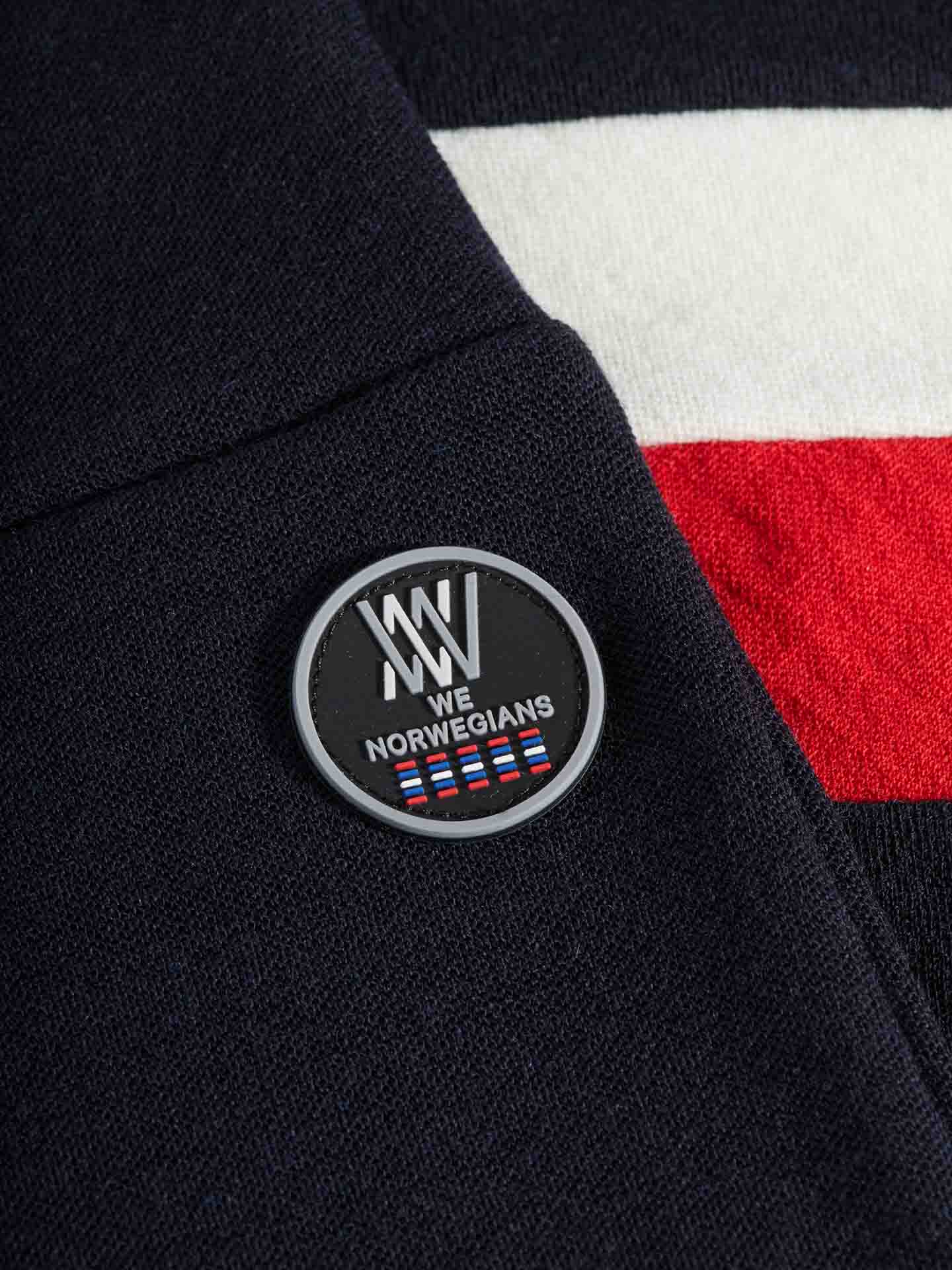 Close-up of a black badge on fabric, featuring the logo "WN We Norwegians" with stylized text and small Norwegian flag icons. The nearby merino wool fabric has a red, white, and blue stripe pattern, making it an ideal base-layer for skiing or complementing your Voss ColBlock Leggings Women by We Norwegians.