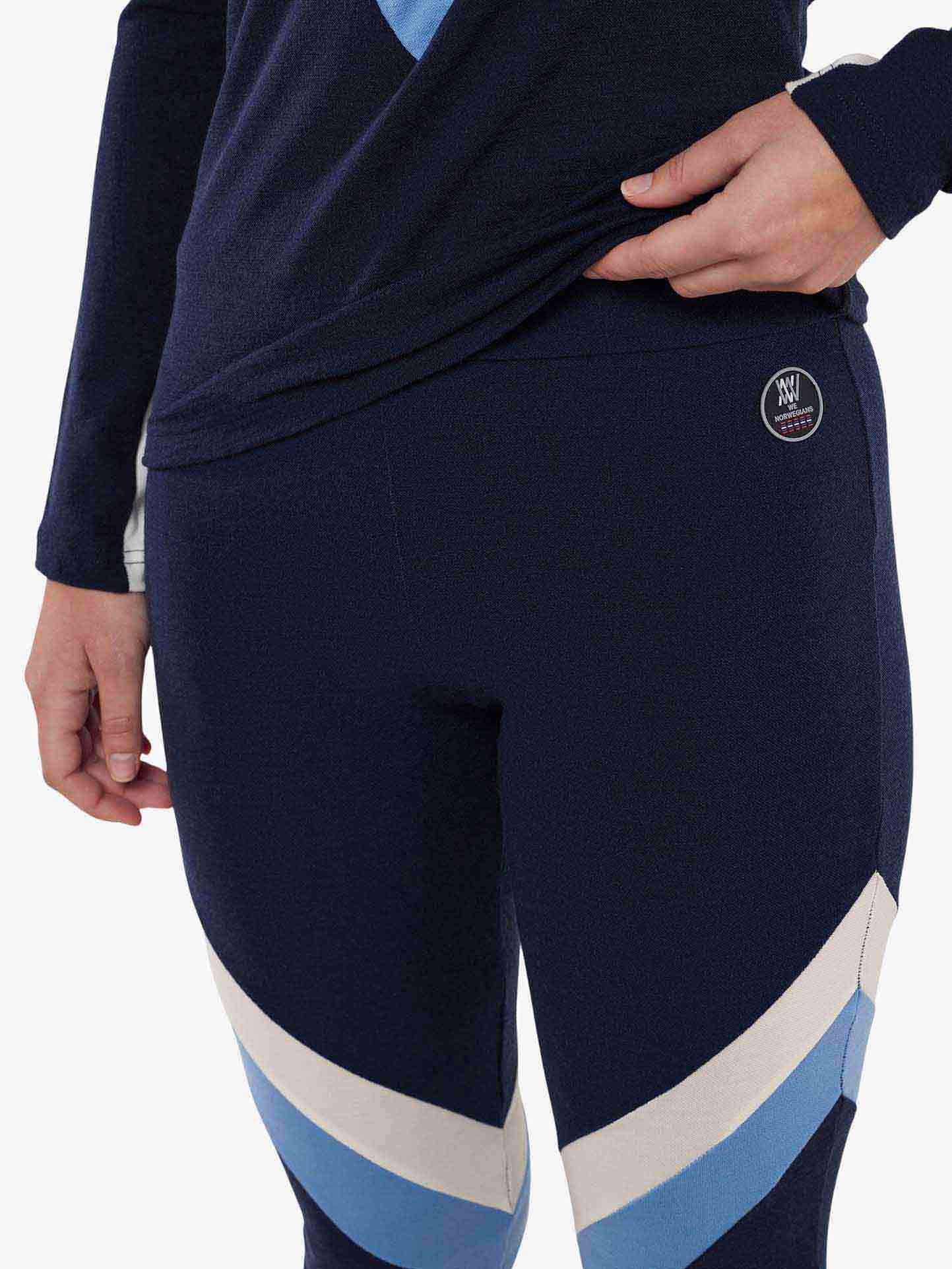 A close-up view of a person wearing Voss ColBlock Leggings Women by We Norwegians, featuring navy blue fabric with light blue and white diagonal stripes near the knees. They are also wearing a long-sleeved navy top made from merino wool. One hand is resting on their hip, making this the perfect base-layer skiing attire.