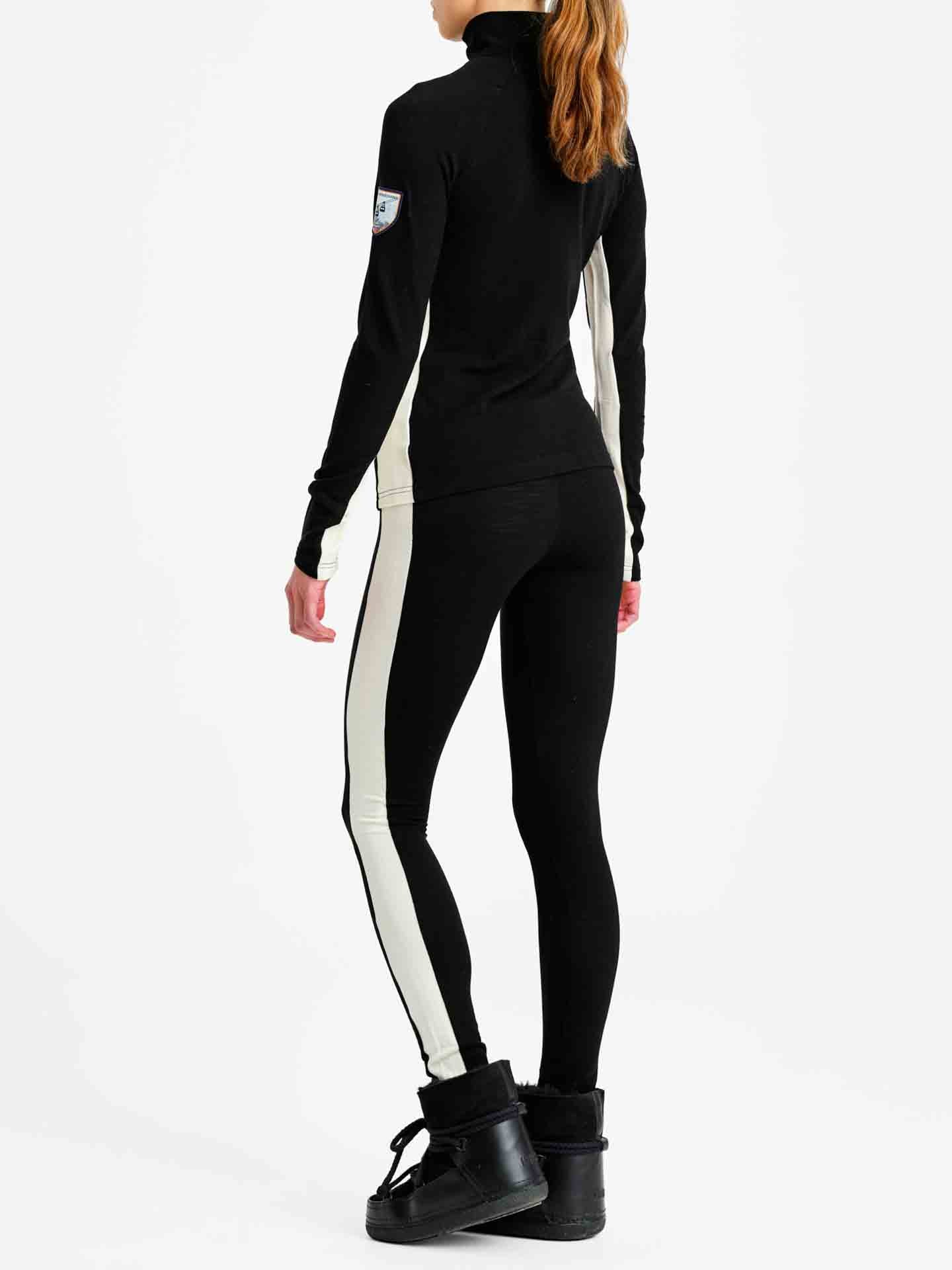 A person stands facing away, dressed in We Norwegians' Voss Leggings for women, paired with a matching high-collared, long-sleeved top. They are wearing black footwear and have their hair pulled back. The race stripe design includes white side stripes on the leggings and a patch on the left sleeve of the top.