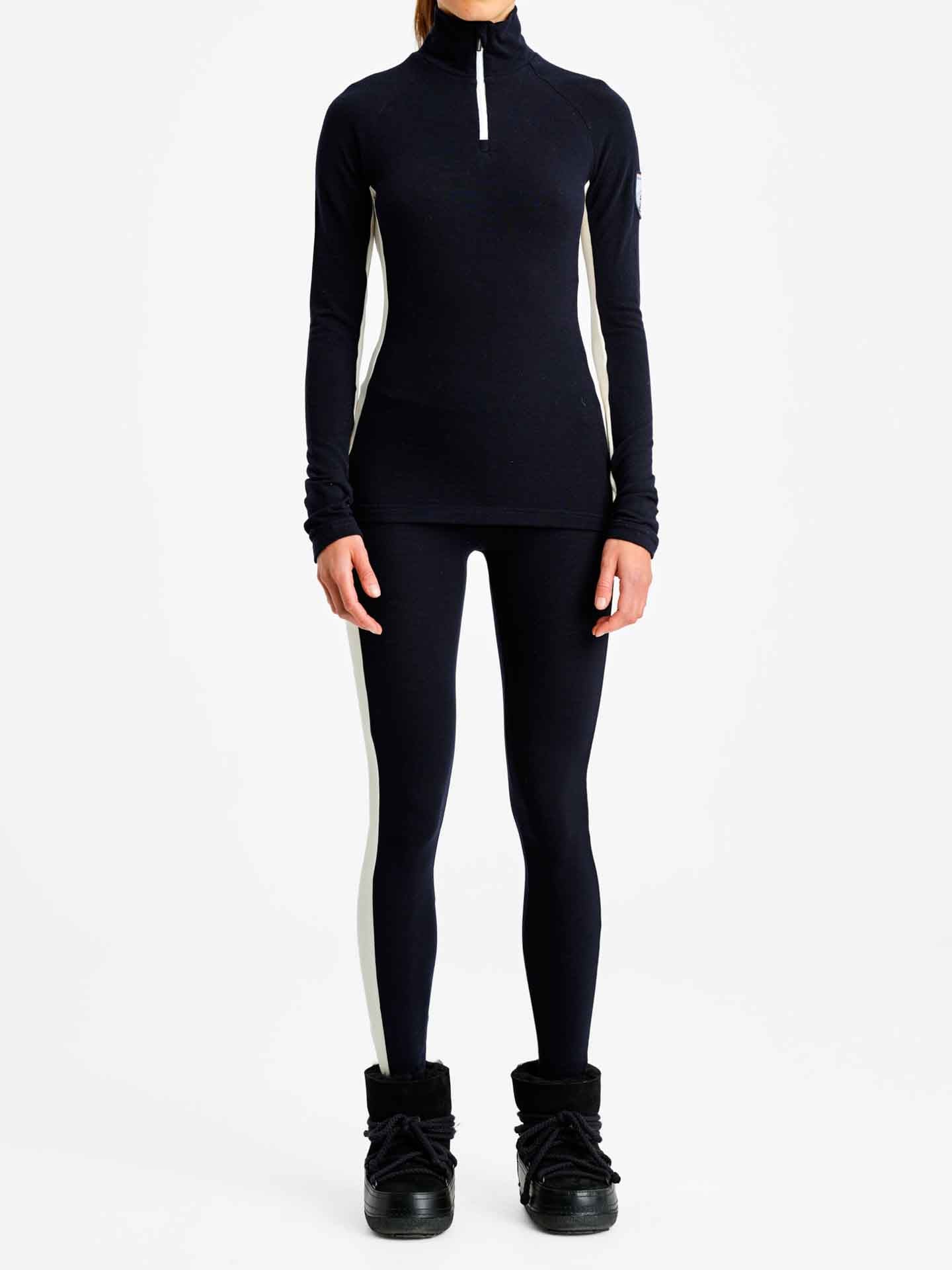 A person standing and facing forward, wearing a black, long-sleeved athletic top with a white race stripe design on the side, We Norwegians Voss Leggings Women featuring matching black base layer leggings with white side panels, and black boots. The background is plain white.