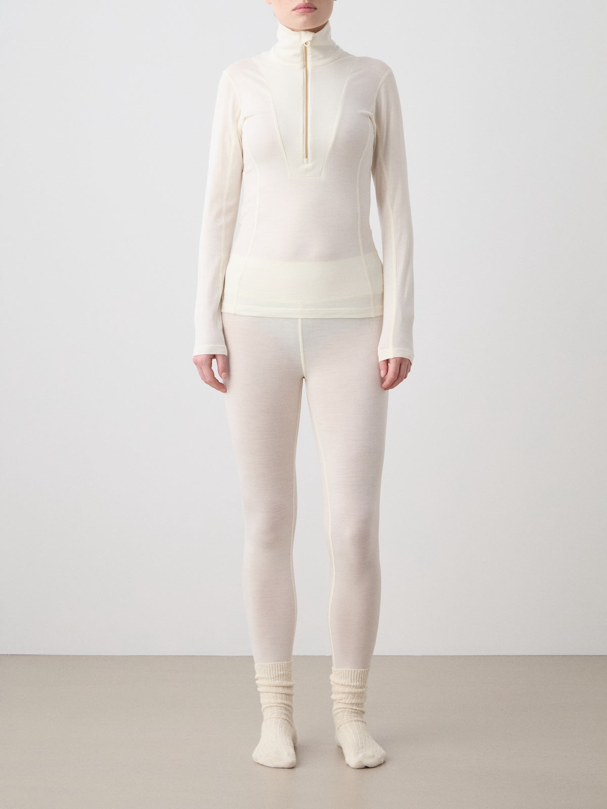 A person stands against a plain background wearing the We Norwegians Sno Merino Zip Up Women—a long-sleeve, off-white zip-up top—paired with matching leggings and socks. Made from breathable single jersey crafted with superfine merino wool, this Scandinavian luxury base layer provides comfort along with understated elegance. Their arms rest at their sides and their face is not shown.