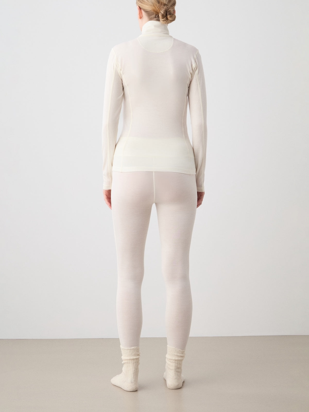 A person stands with their back to the camera, wearing the We Norwegians Sno Merino Zip Up Women—a fitted, long-sleeve white top—paired with matching white leggings crafted from breathable jersey fabric and cream-colored socks, set against a plain neutral background.