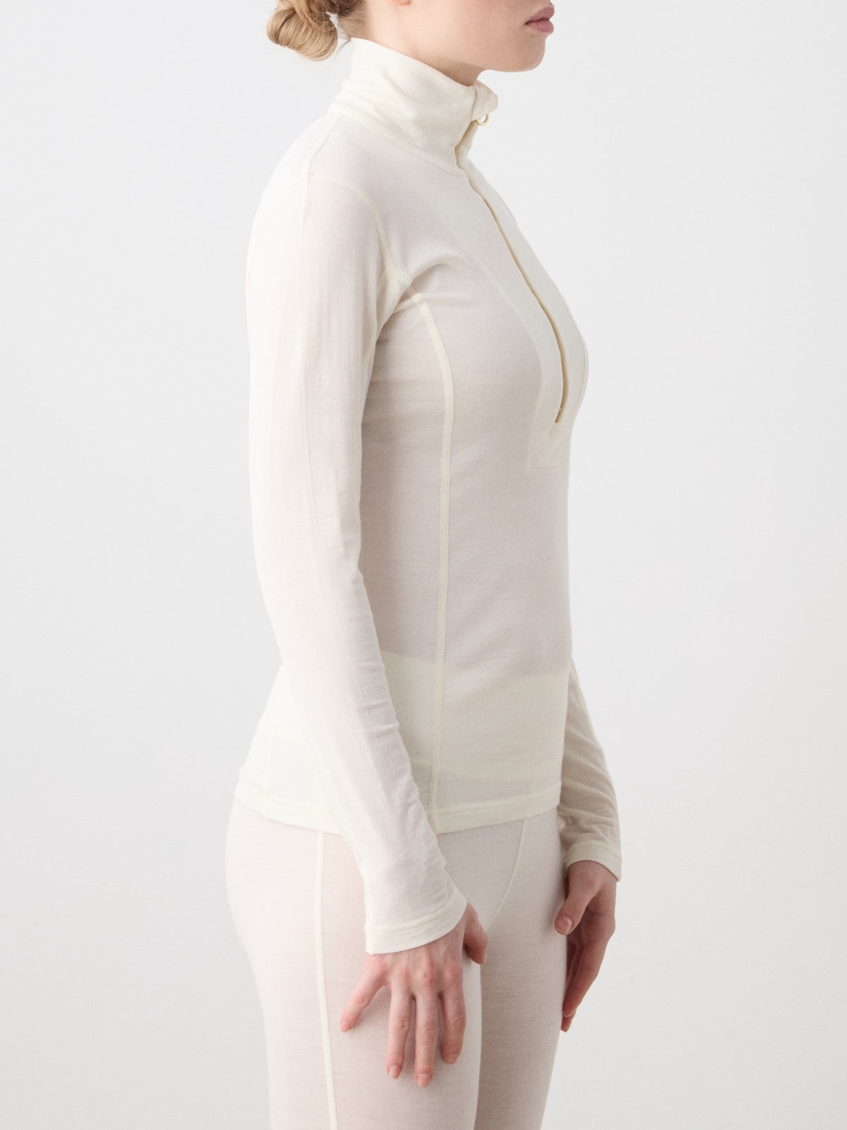 A woman stands in profile against a plain white background, embodying Scandinavian luxury base layer style while wearing the We Norwegians Sno Merino Zip Up Women—a fitted, long-sleeve, cream-colored top and matching bottoms crafted from superfine merino wool.