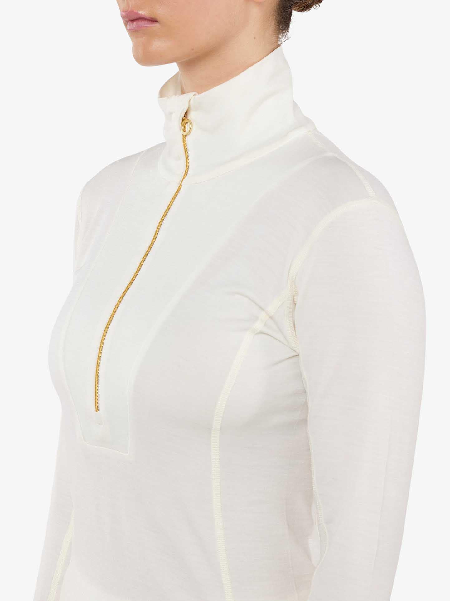 A person dressed in the We Norwegians Sno Merino Zip Up Women, a close-fitting, long-sleeved, off-white top with a high collar and a gold vertical half-zip closure made from super fine merino wool, highlighting its Scandinavian luxury base-layers. Their hair is neatly pulled back, and the background is plain white.