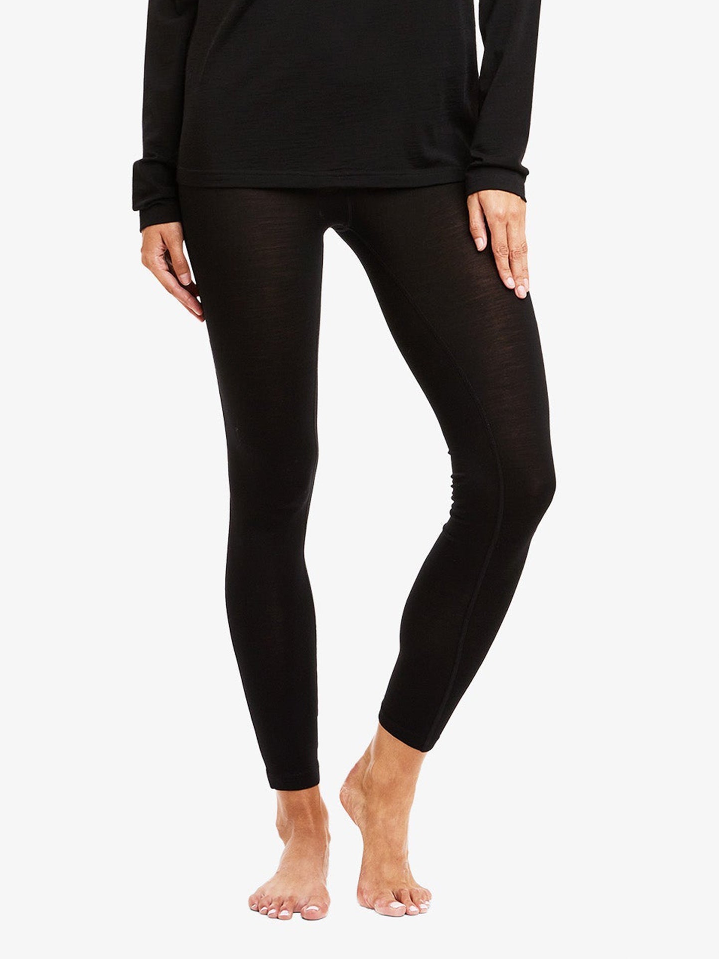 A person is standing barefoot with feet slightly apart, wearing black, fitted Sno Merino Leggings Women by We Norwegians and a long-sleeve black top made of super fine merino wool. The image is cropped to show from the shoulders down to the feet. The background is plain white, highlighting the Scandinavian luxury base-layers.