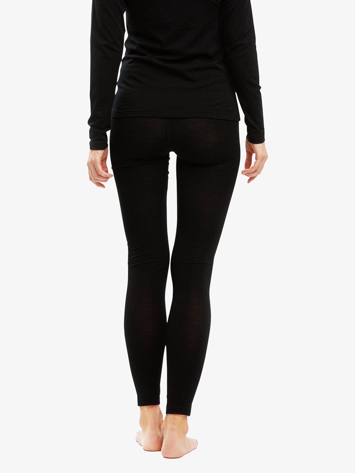 A person is standing with their back facing the camera, wearing a black long-sleeve top and a pair of We Norwegians' Sno Merino Leggings for women, made from super fine merino wool. They are barefoot against a white background.