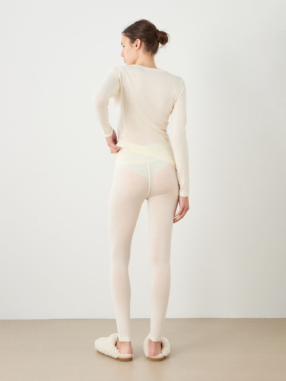 A woman stands facing away, wearing a long-sleeve cream top, We Norwegians Sno Merino Leggings for women in a matching shade, white breathable wool underwear, and fuzzy slippers, her hair styled in a bun against a plain background.