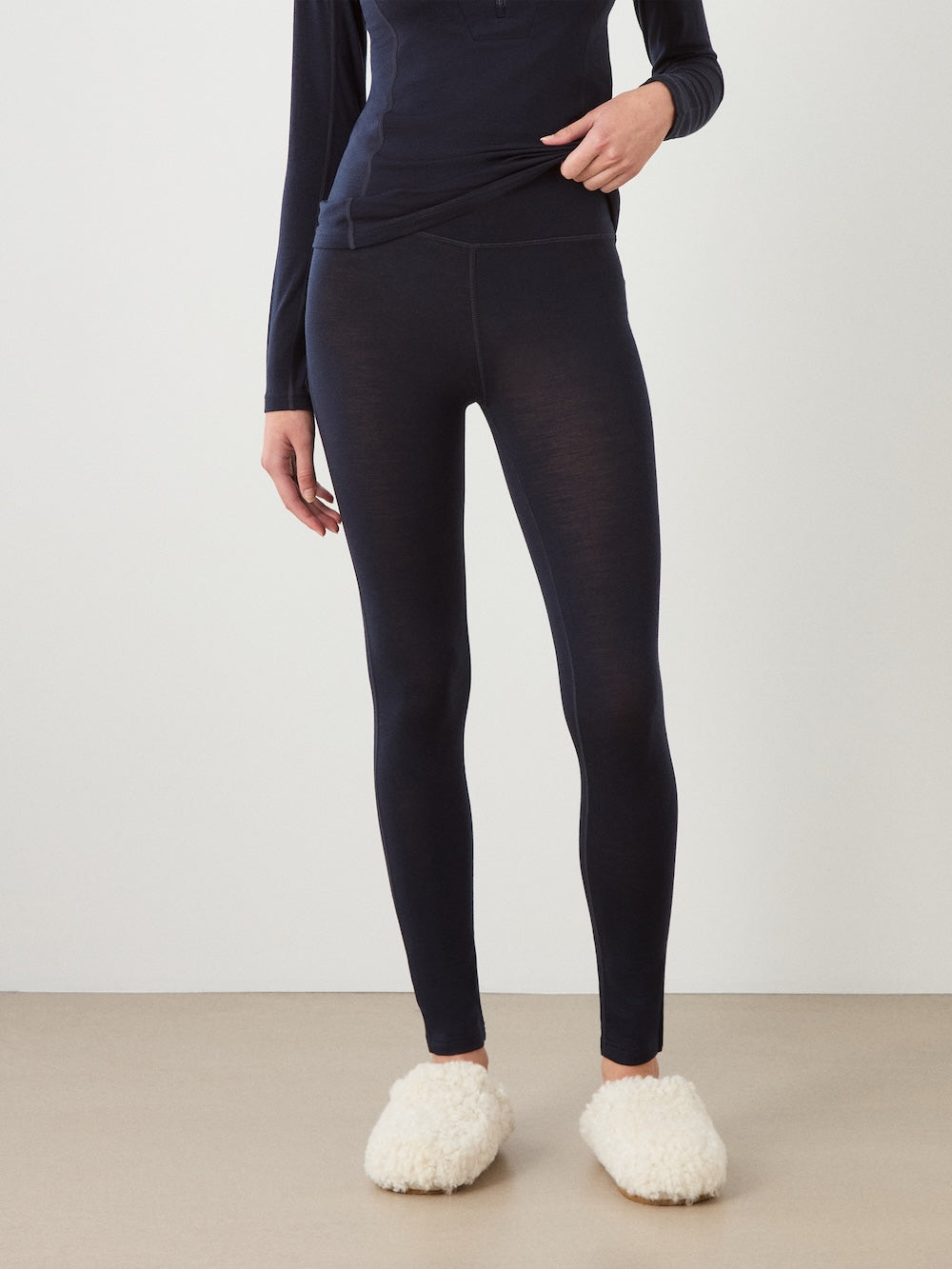 A person stands indoors against a plain light wall, wearing We Norwegians Sno Merino Leggings for women in dark navy, paired with a matching long-sleeve top and white fuzzy slippers. Only the body from shoulders down is visible, with hands relaxed by their sides.