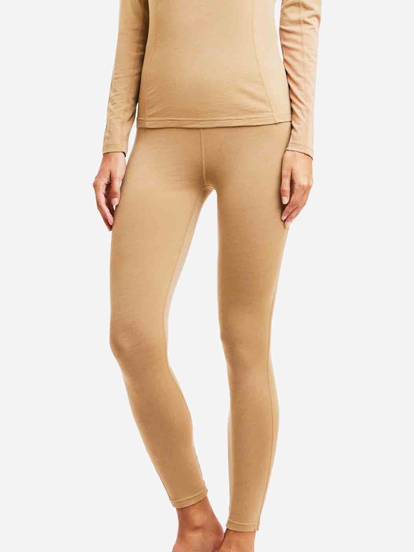 A partial view of a person standing against a plain white background, showcasing the bottom half of their body dressed in form-fitting beige *Sno Merino Leggings Women* by *We Norwegians*. The leggings, crafted from super fine merino wool, appear smooth and stretchy.